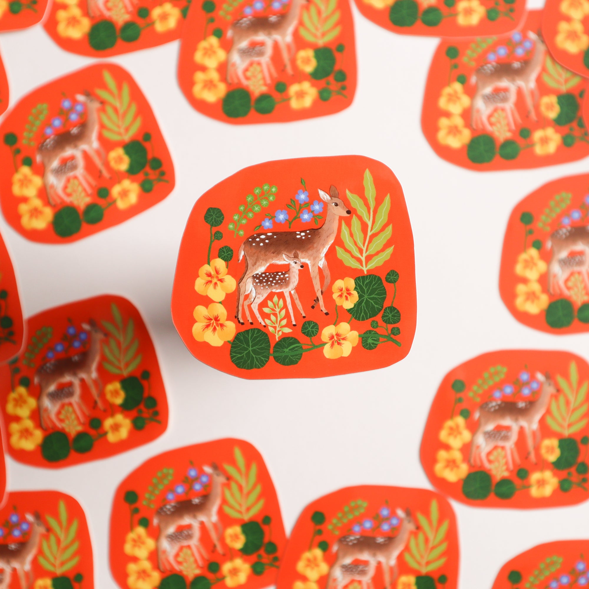 The Deer & Fawn Sticker by Papio Press shows a deer and her faun amid flowers and leaves on an orange background, held up above identical UV-protected stickers scattered on a white surface.
