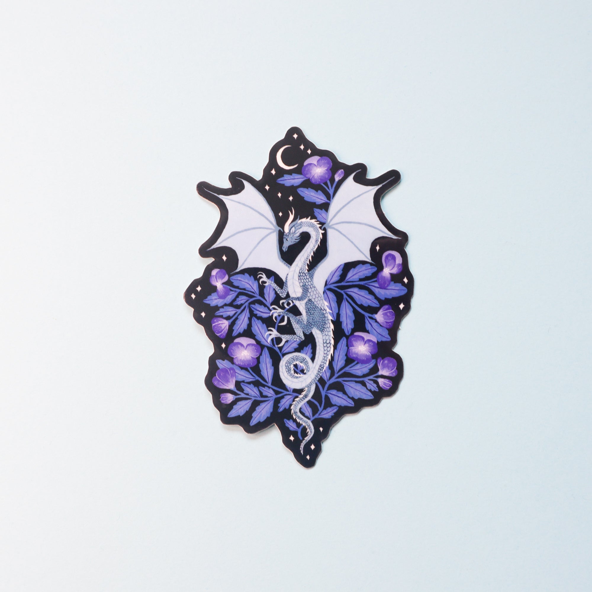 From the Papio Press Fantasy Sticker Collection, this glossy vinyl sticker shows a white dragon with wings spread among purple flowers and green leaves, crescent moon above, and includes UV protection. Set on a pale blue background.