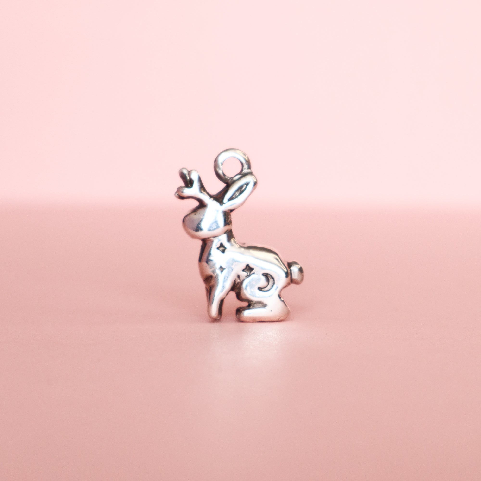 The Silver Lunar Jackalope Pendant by Papio Press is a small, shiny recycled silver pendant shaped like a sitting jackalope with antlers, shown against a soft pink background.