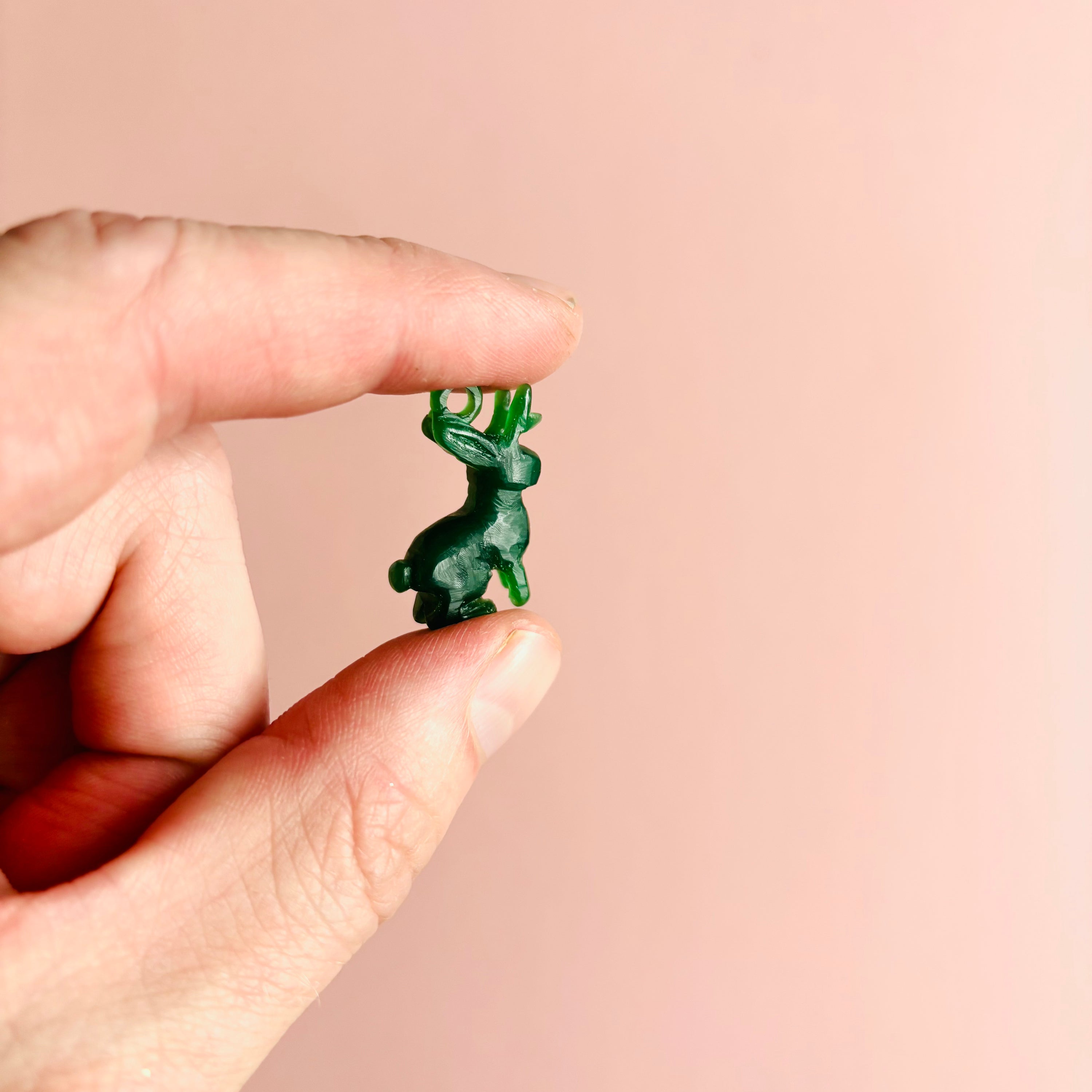 A hand holds the Lunar Jackalope's original green wax carving against a pale pink background.