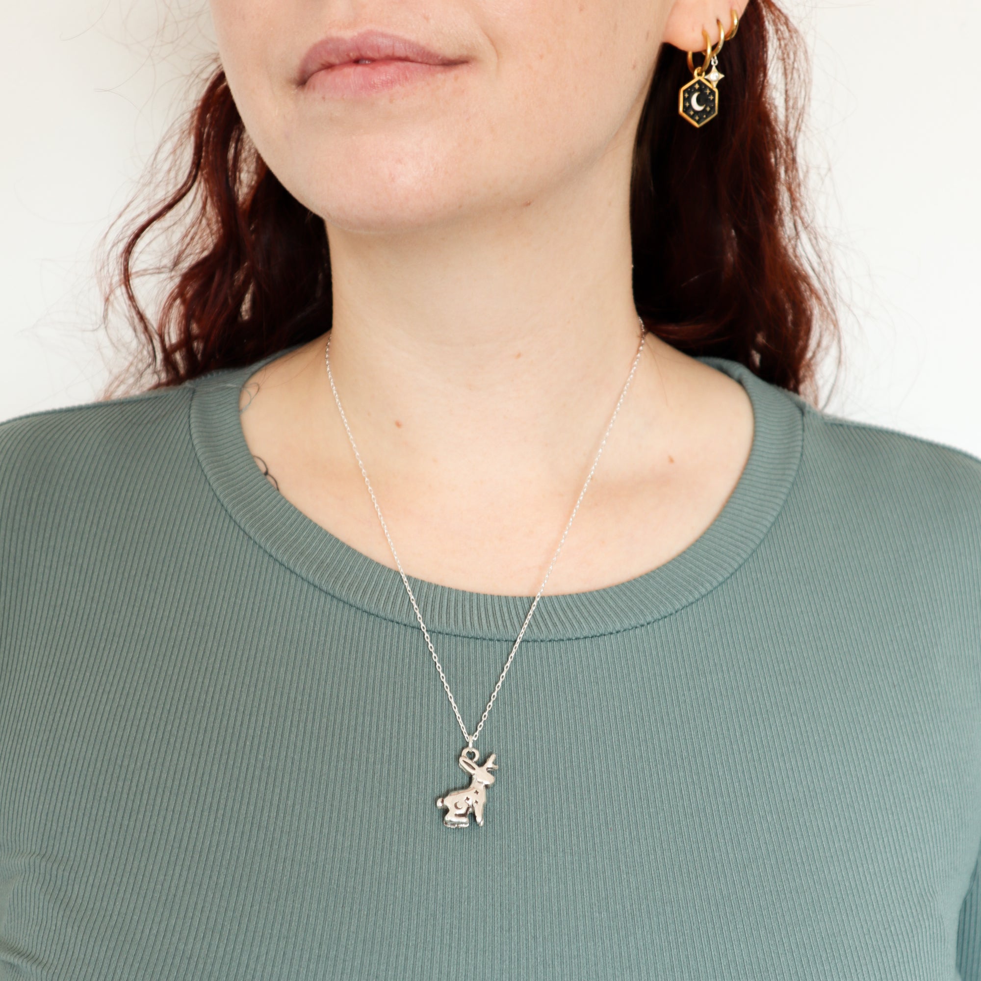 A woman stands against a plain white background, wearing a ribbed light green top, Papio Press's handmade Silver Lunar Jackalope Pendant necklace.