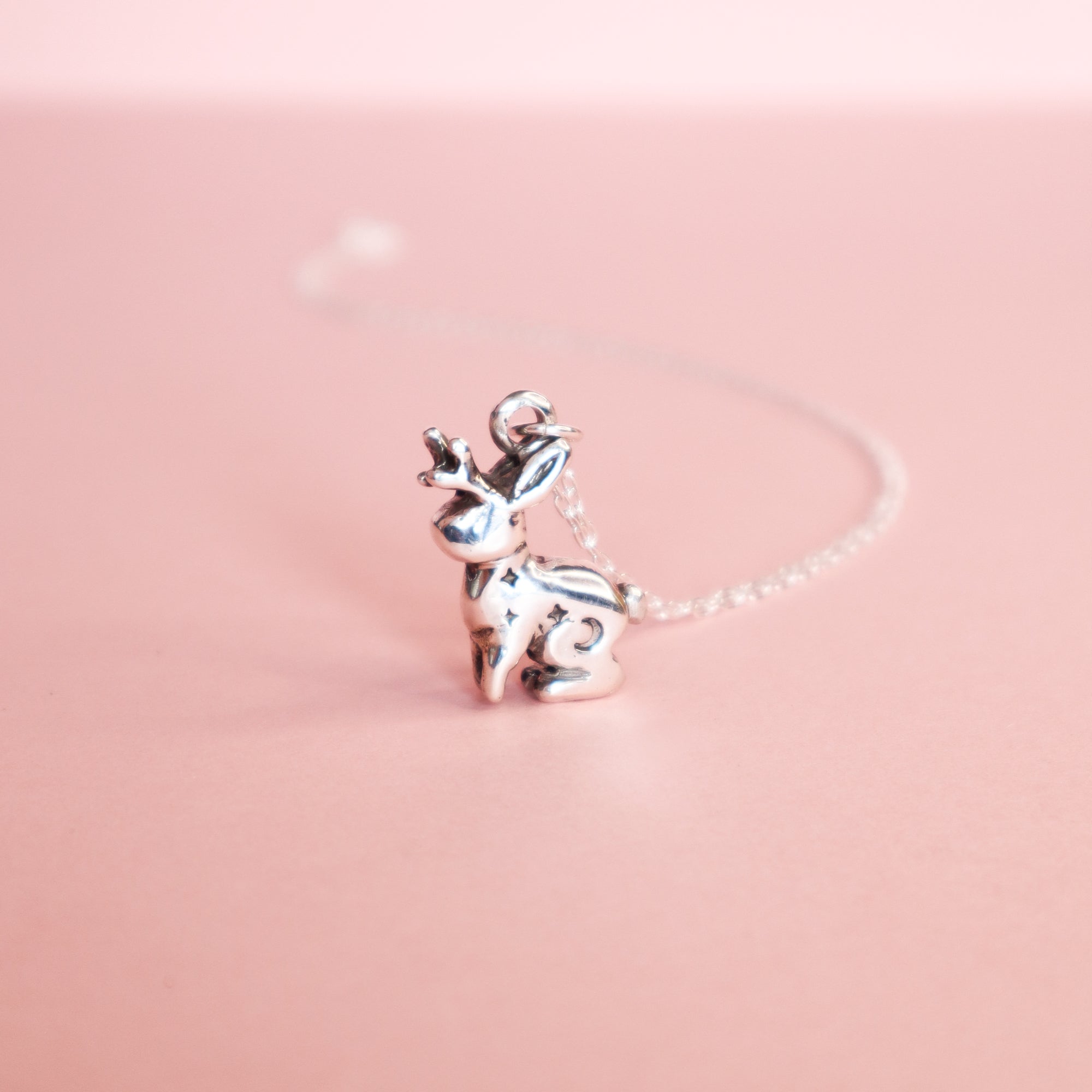 The Silver Lunar Jackalope Pendant by Papio Press, a handmade silver jewelry piece, sits on a pastel pink surface with a softly blurred pink background.