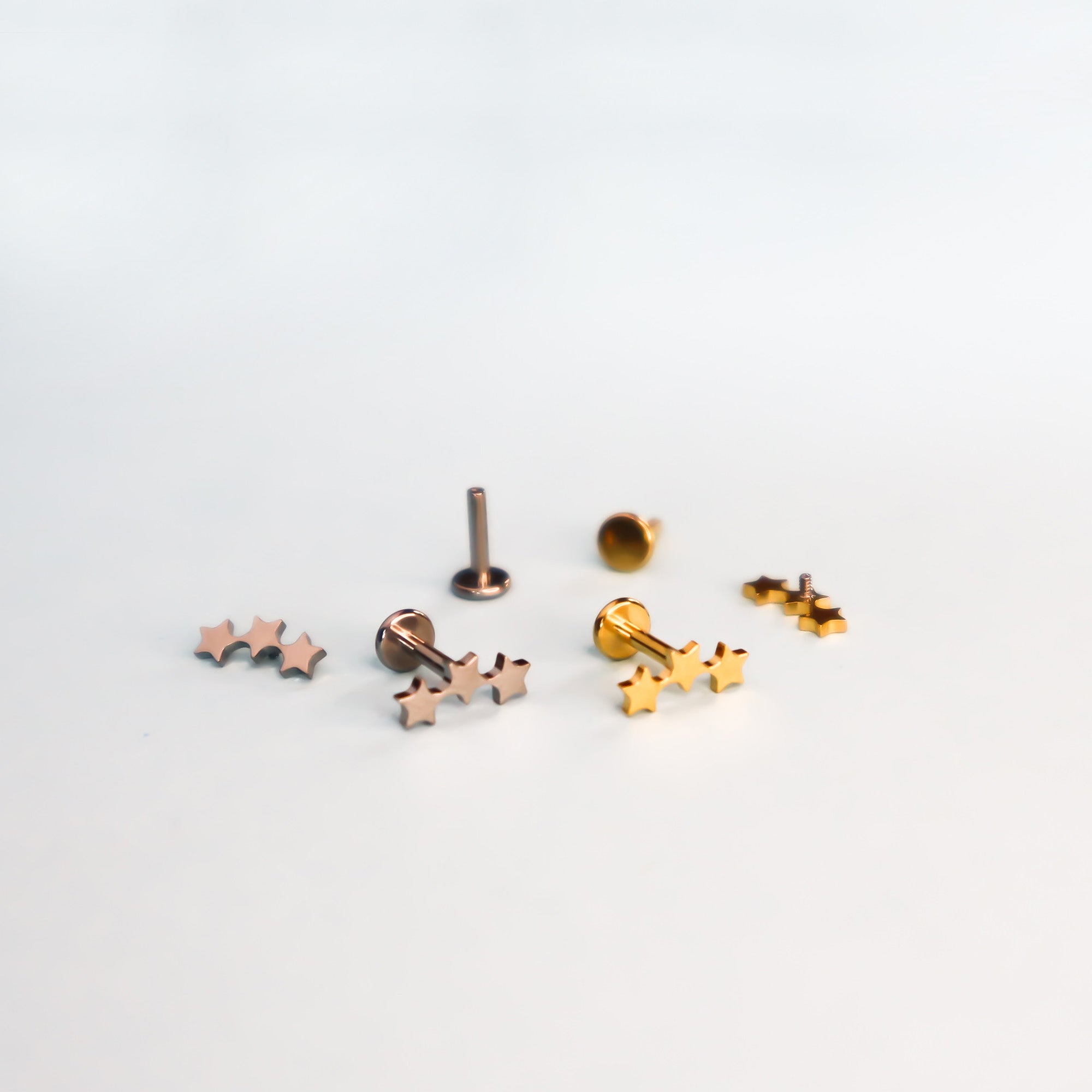The Papio Press Star Trio Labret Earring features a titanium base in gold and rose gold tones, perfect for cartilage piercings. Displayed on a white surface, some earrings lay flat while others stand upright to show their backing posts.