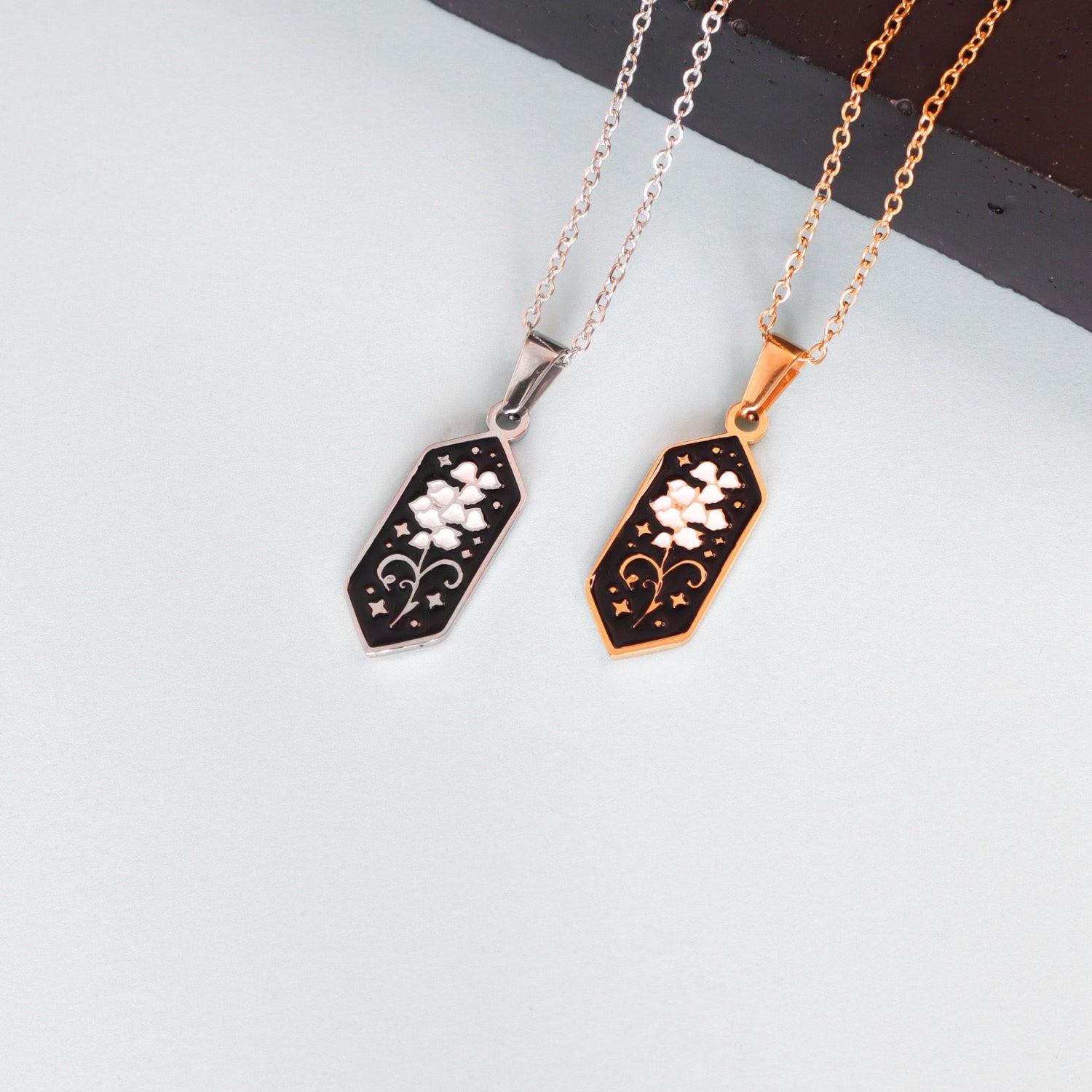 Two Papio Press Lily of the Valley Pendant Enamel Necklaces, each with a hexagonal pendant featuring a white flower on black; one with a silver chain and frame, the other with an 18K gold-plated chain and frame, both displayed on a light surface.
