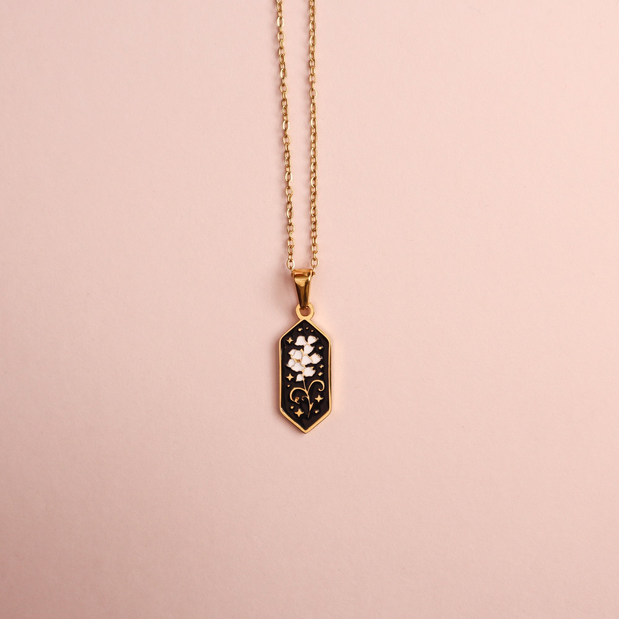 The Lily of the Valley Pendant Enamel Necklace by Papio Press features a gold hexagonal pendant with a black background and delicate white floral design, beautifully displayed on a light pink surface.
