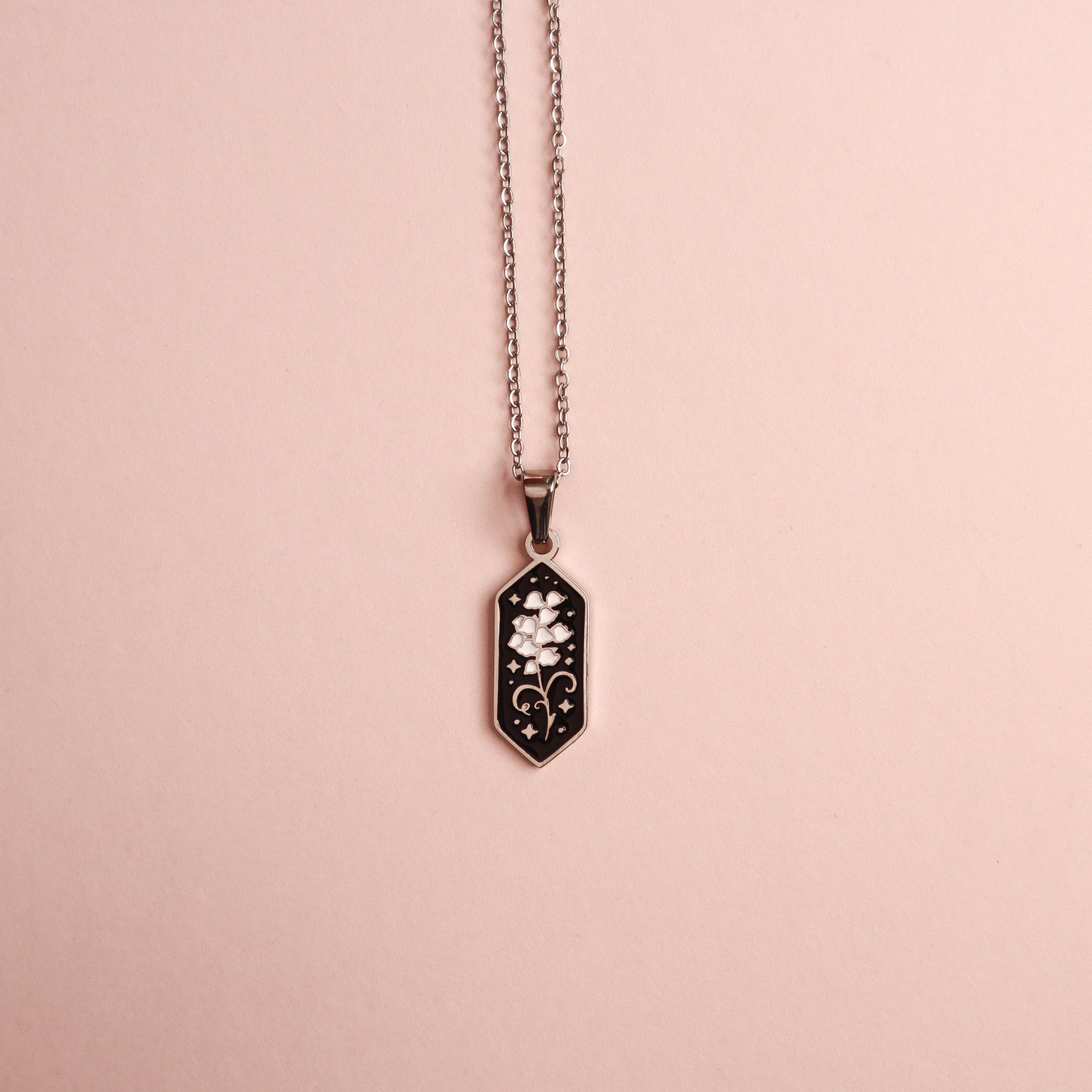 The Lily of the Valley Pendant Enamel Necklace by Papio Press features a hexagonal silver and 18K gold plated pendant with a floral decorative design, shown on a light pink background.