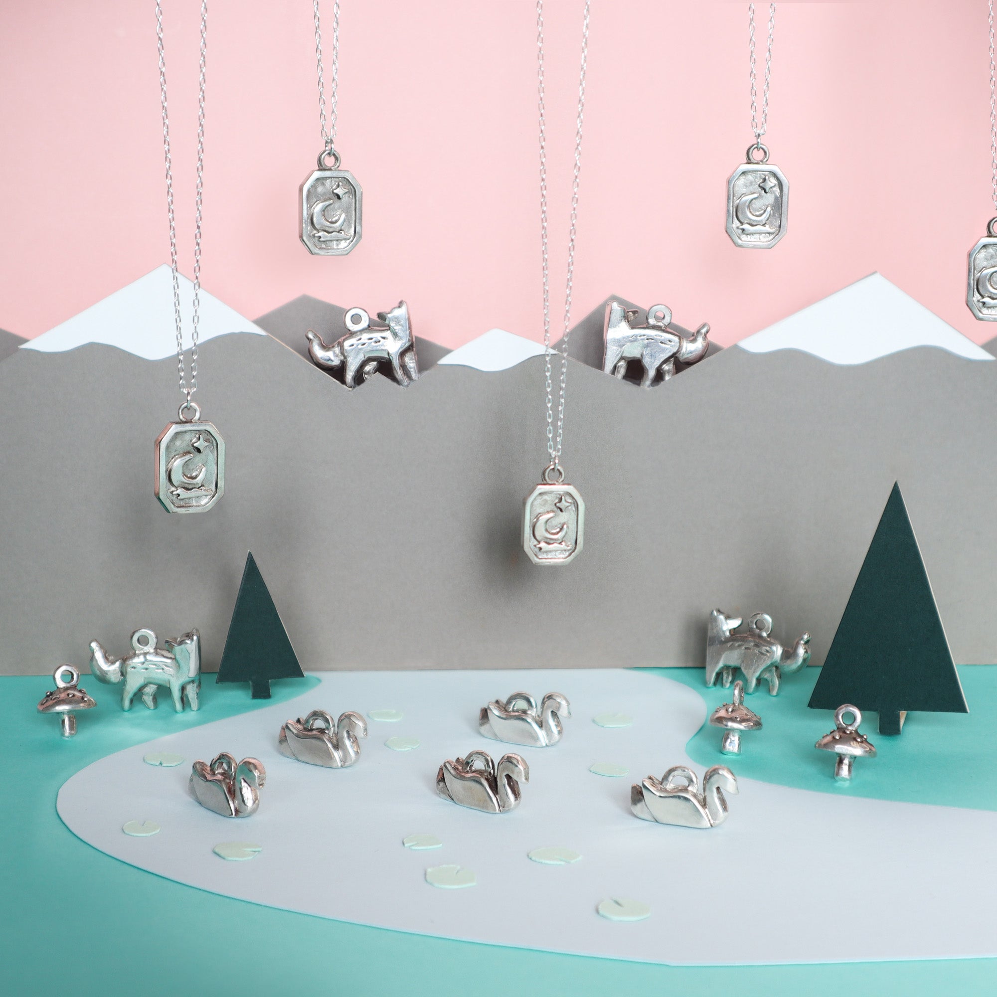 Papio Presss handmade Silver Mushroom Pendant hangs above a paper landscape of mountains, trees, and a pond, while matching recycled silver mushroom pendants and figurines are artfully arranged on the ground and water.