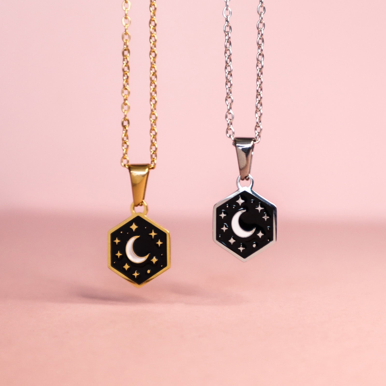 Papio Press Mini Hexagon Moon Pendants feature gold and silver stainless steel necklaces with crescent moon and star designs on black enamel, each on matching chains, shown against a pink backdrop.