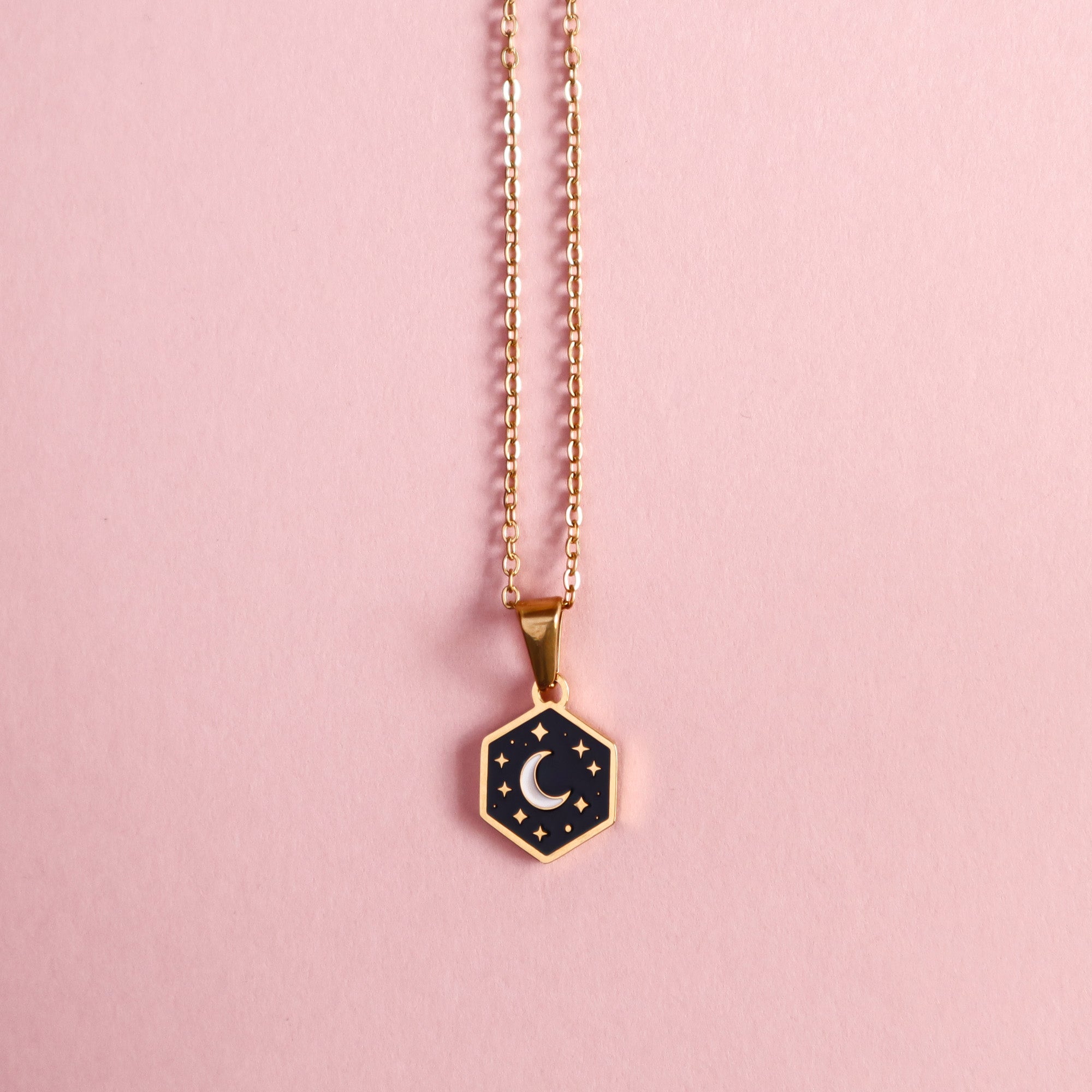The Papio Press Mini Hexagon Moon Pendant is a gold stainless steel necklace with a black enamel center featuring a crescent moon and small stars, shown on a light pink background.