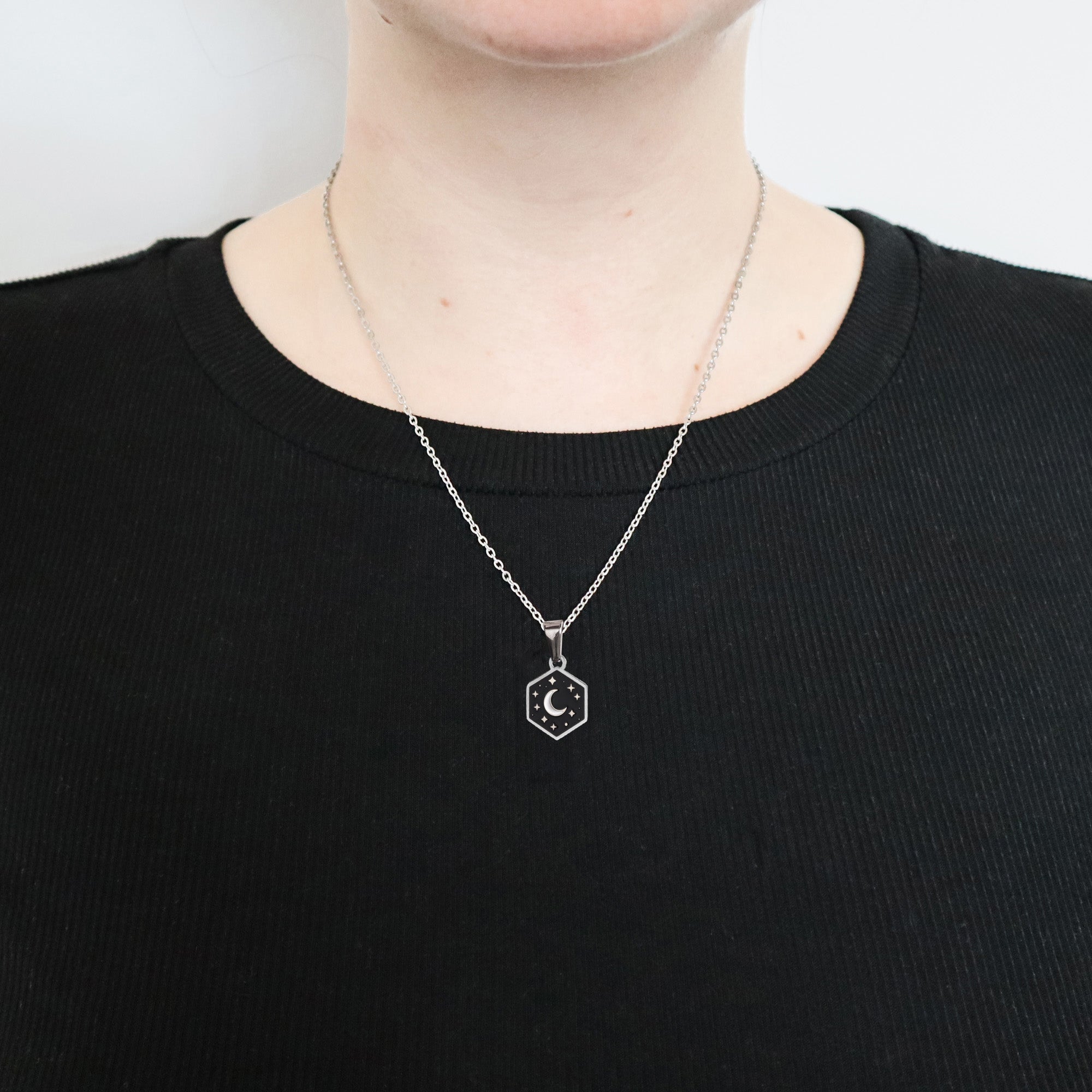 A person wears a black ribbed shirt and the Papio Press Mini Hexagon Moon Pendant, a silver chain enamel necklace with a crescent moon and star design. Only the lower face and neck are visible.
