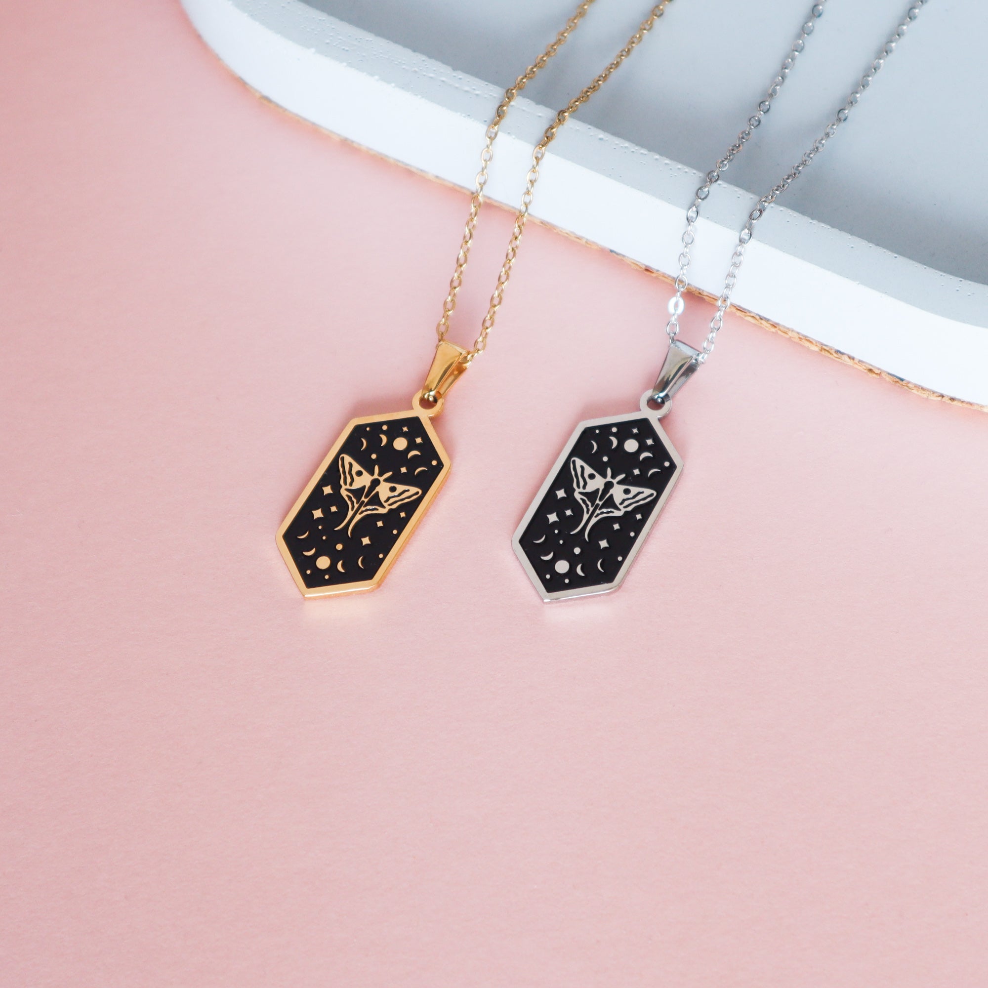 Two Papio Press Moth Pendant Enamel Necklaces, one gold plated and one silver, each with an engraved floral and crescent moon design, are displayed on a light pink surface with a white edge in the background.