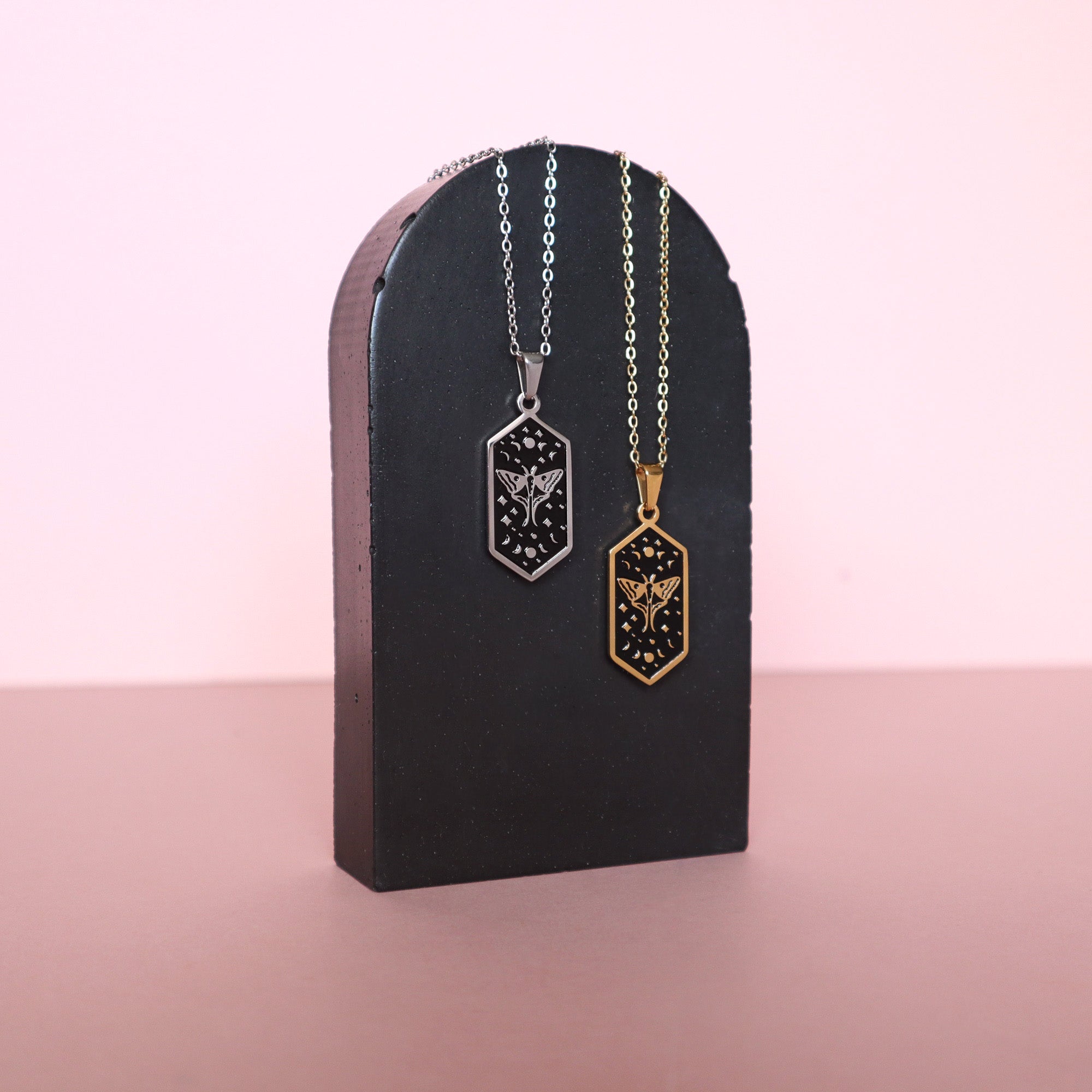 Papio Press’s Moth Pendant Enamel Necklaces, in gold- and silver-plated finishes, feature delicate moth designs on hexagonal pendants. Shown on a black arch stand with a pink backdrop, they offer an elegant, modern display.