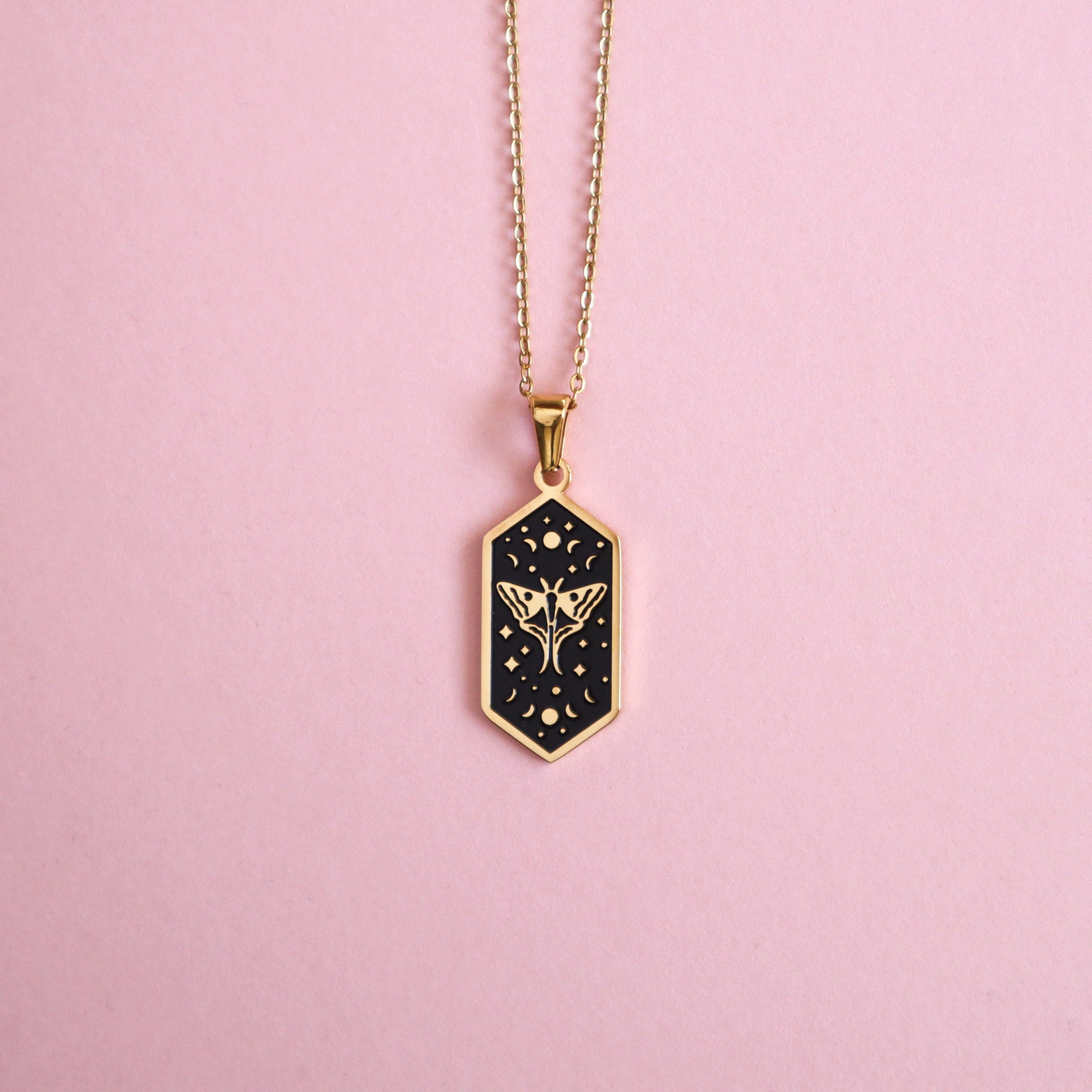 The Moth Pendant Enamel Necklace by Papio Press features a hexagonal gold-plated black pendant with a delicate gold moth and floral design, displayed on a light pink background.