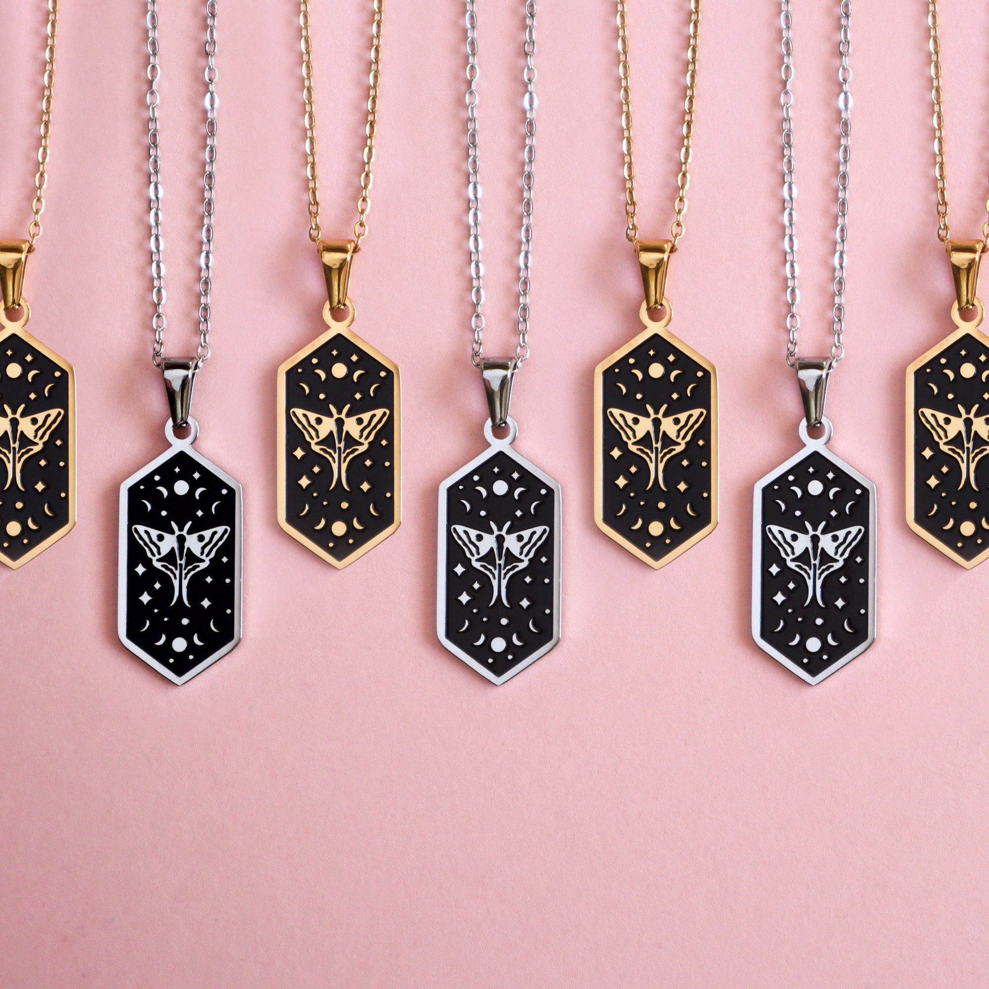 Papio Press Moth Pendant Enamel Necklaces feature hexagonal black pendants with gold or silver floral and star designs, each on alternating gold plated or silver chains, elegantly displayed in a row against a pink background.
