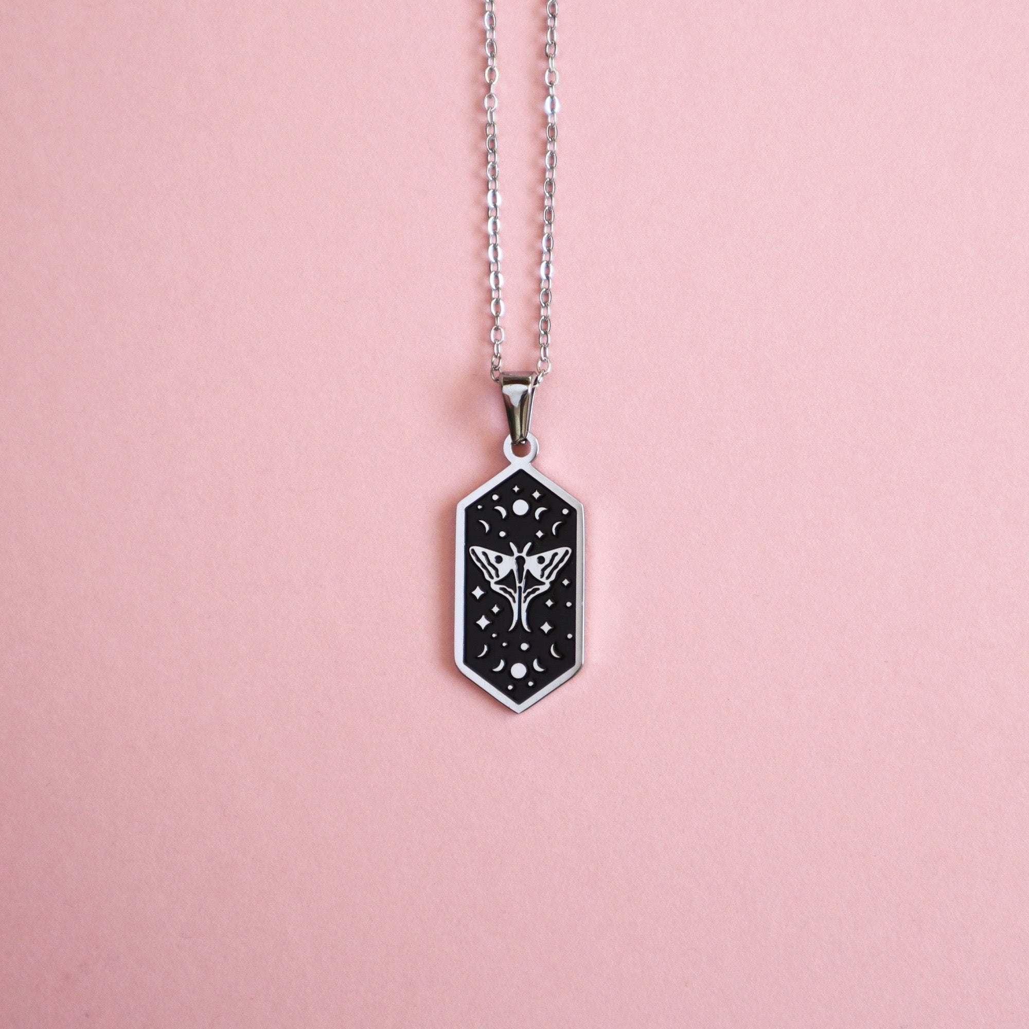 The Moth Pendant Enamel Necklace by Papio Press is a silver necklace with a hexagonal black pendant featuring a white floral and star design, shown on a light pink background.