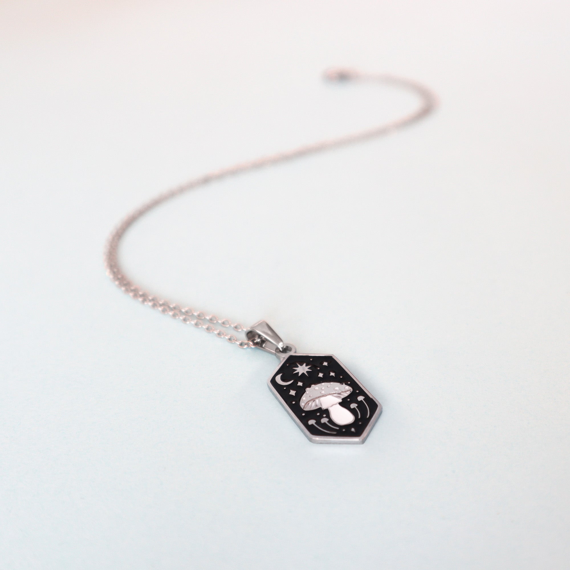 The Papio Press Mushroom Pendant Enamel Necklace features a hexagonal black pendant with engraved mushrooms, stars, and a crescent moon, shown on a light blue background.