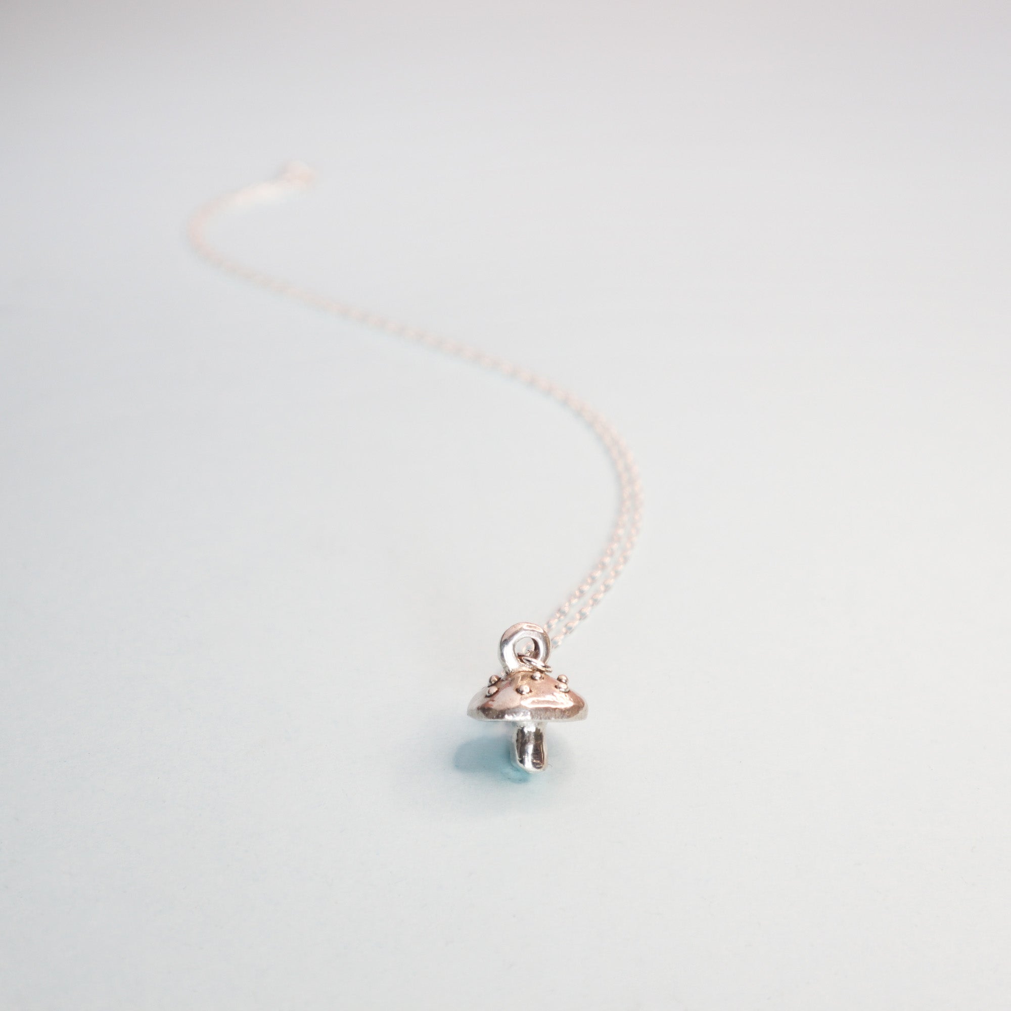 Papio Press Silver Mushroom Pendant: A delicate handmade silver necklace featuring a small mushroom-shaped recycled silver pendant on a soft blue surface. An extended chain adds depth and enhances its minimalist design.