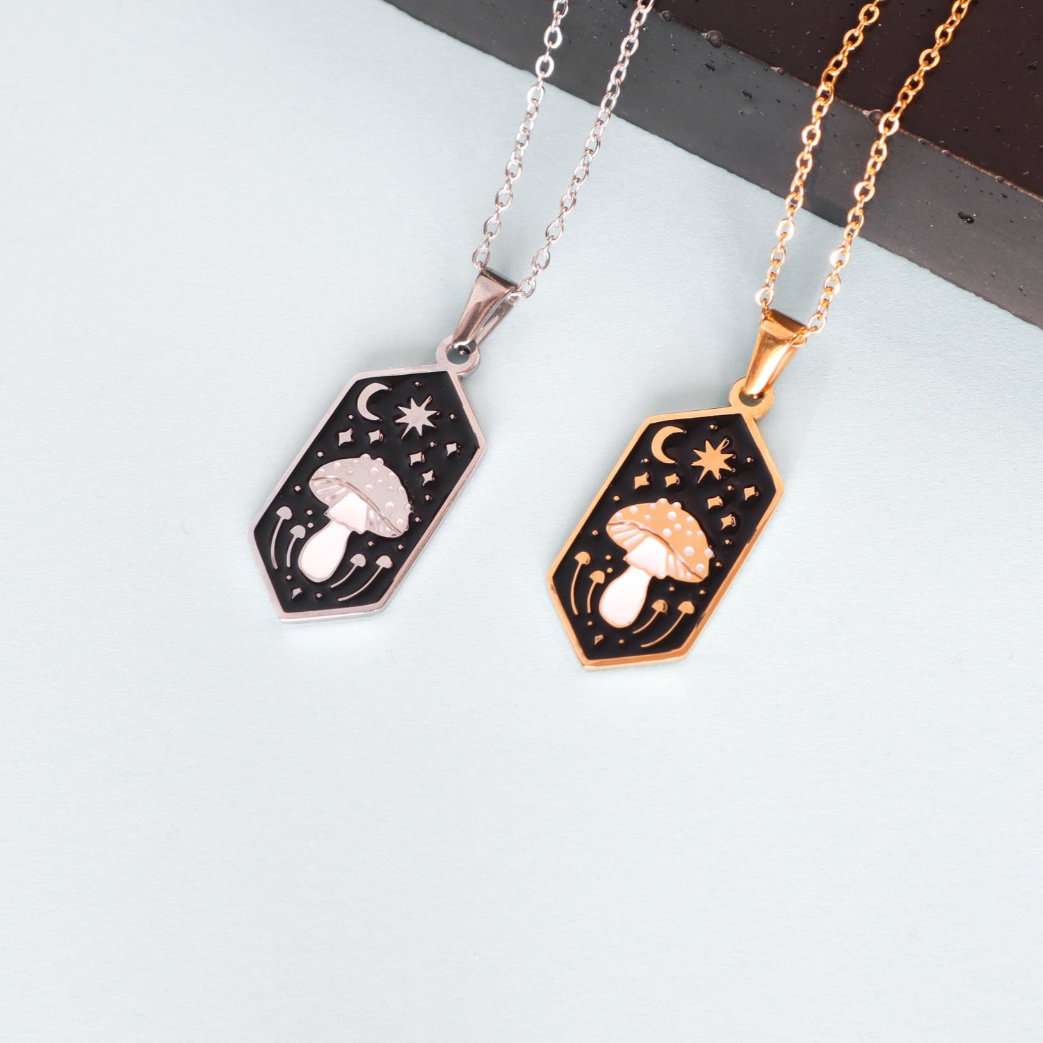 Two Papio Press Mushroom Pendant Enamel Necklaces—one silver and one 18K gold plated—feature etched mushroom, crescent moon, and star designs on black enamel pendants, displayed on a light surface.