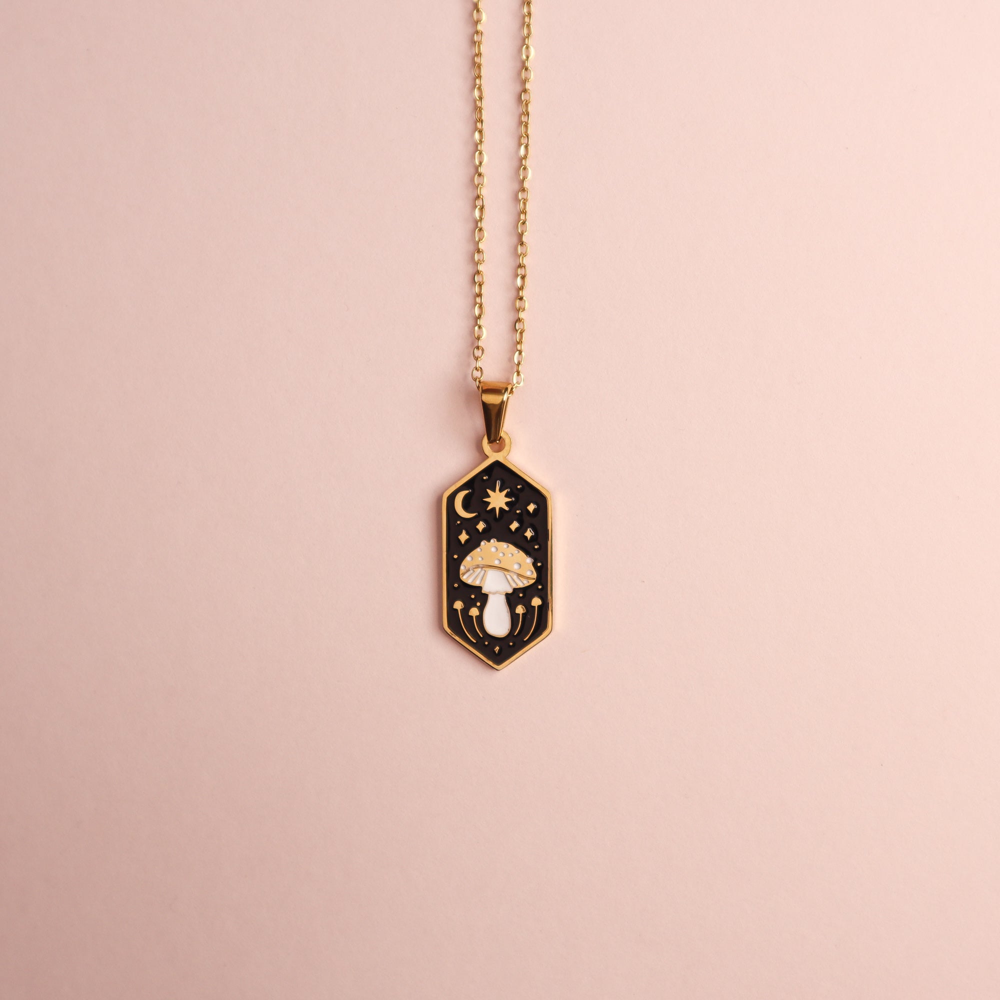 The Papio Press Mushroom Pendant Enamel Necklace features an 18K gold-plated chain and a hexagonal black enamel pendant with a white mushroom, stars, and crescent moon, shown against a light pink background.