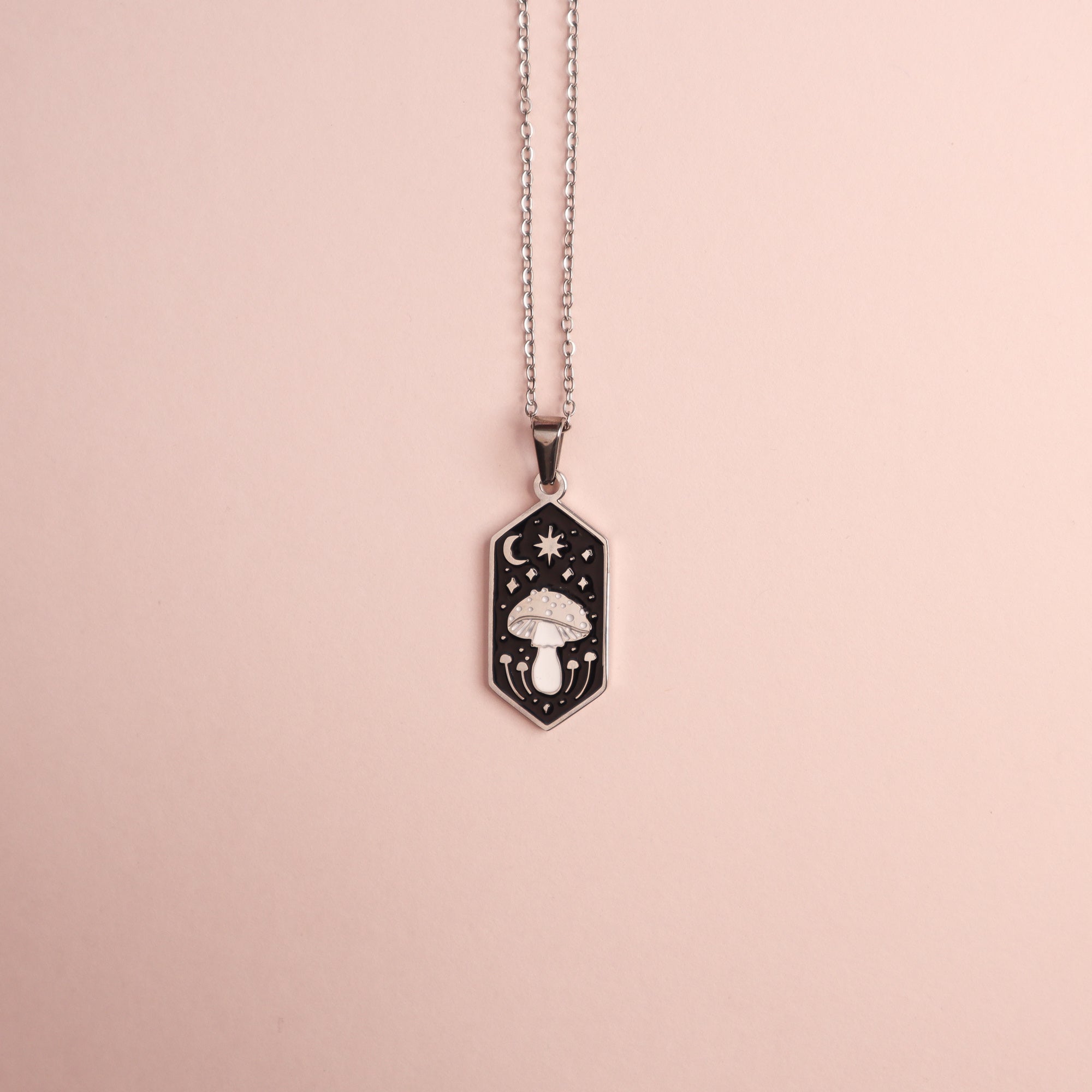 The Papio Press Mushroom Pendant Enamel Necklace features a silver hexagonal pendant with an engraved mushroom, stars, and crescent moon on a black enamel background, displayed on a pale pink surface.