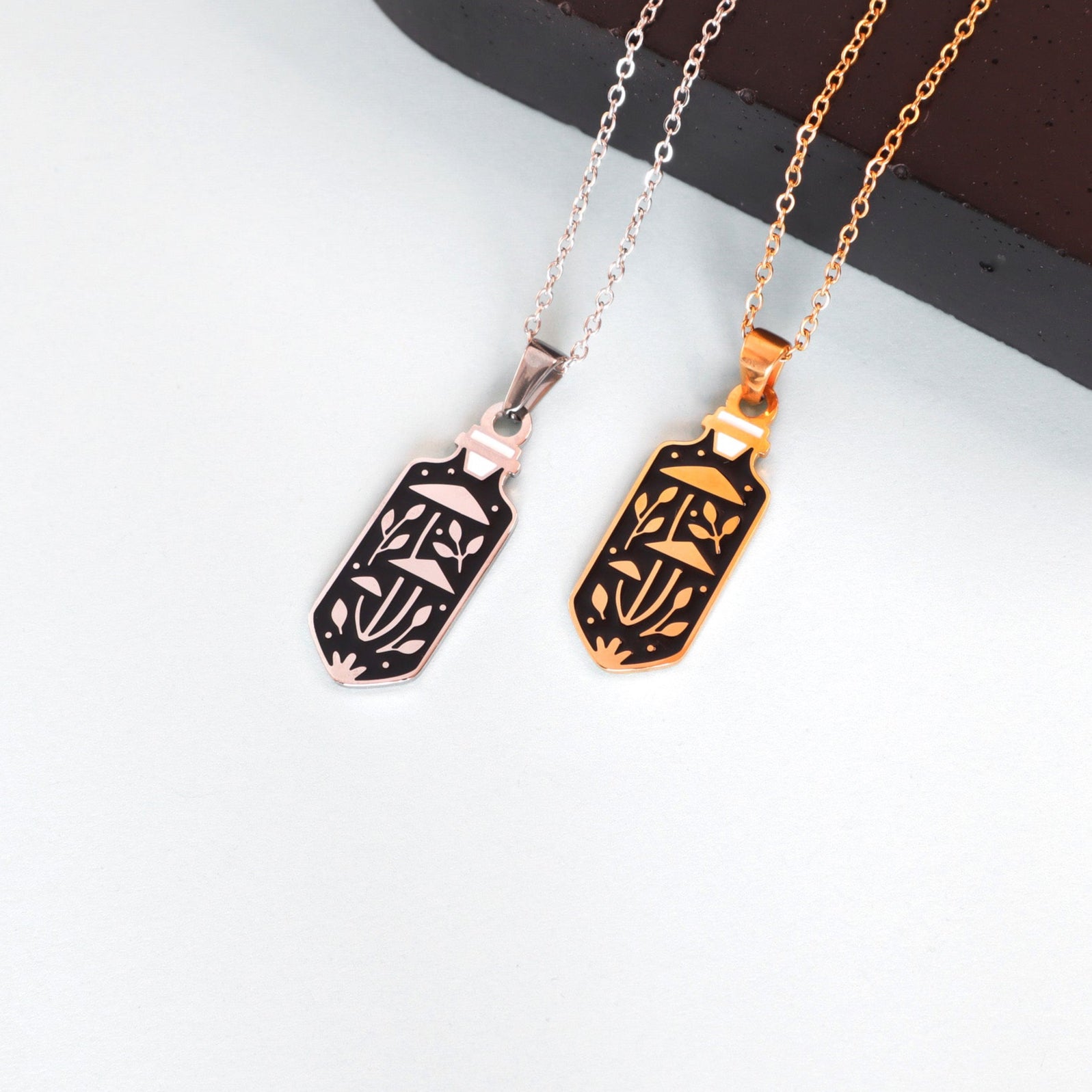 Two Mushroom Potion Pendant Enamel Necklaces by Papio Press—one gold plated, one silver—each features an intricate rectangular line art design. Both are shown on a light surface with a dark brown object behind them.