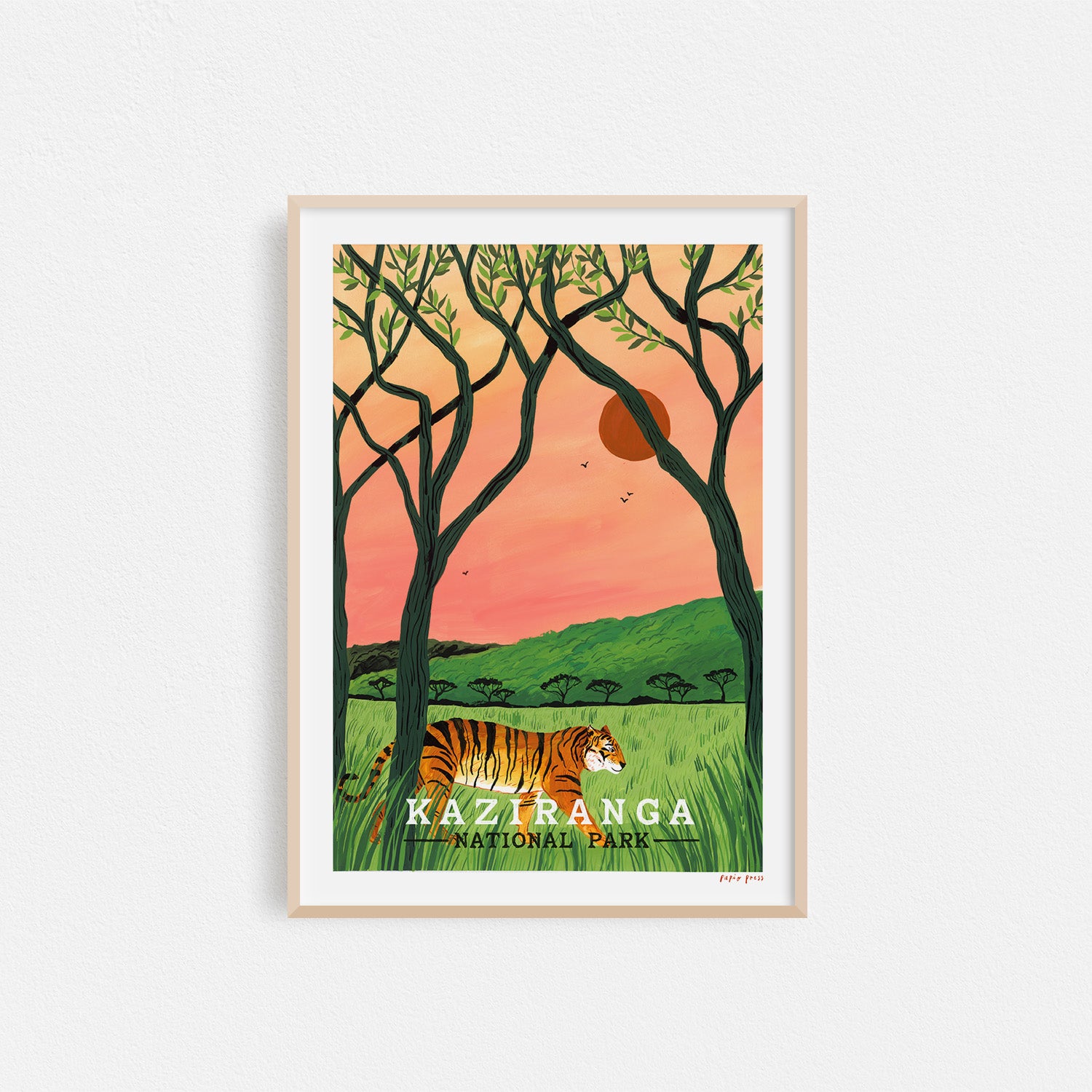 The Kaziranga National Park Artists Print by Papio Press features a tiger walking through tall grass with trees and birds against a pink-orange sky and green hills, inkjet printed on archival satin paper.