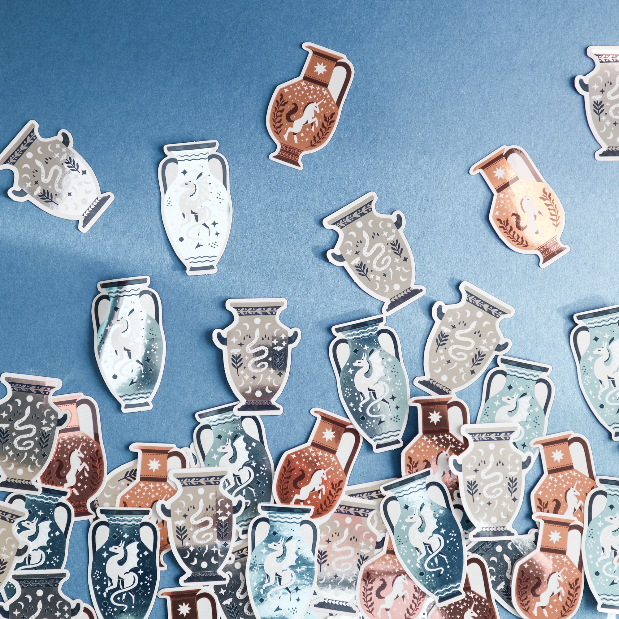 Papio Press Unicorn Vase Stickers are scattered on a blue surface, featuring illustrated vases with vibrant colors, copper mirror metallic finishes, and decorative motifs inspired by ancient pottery.