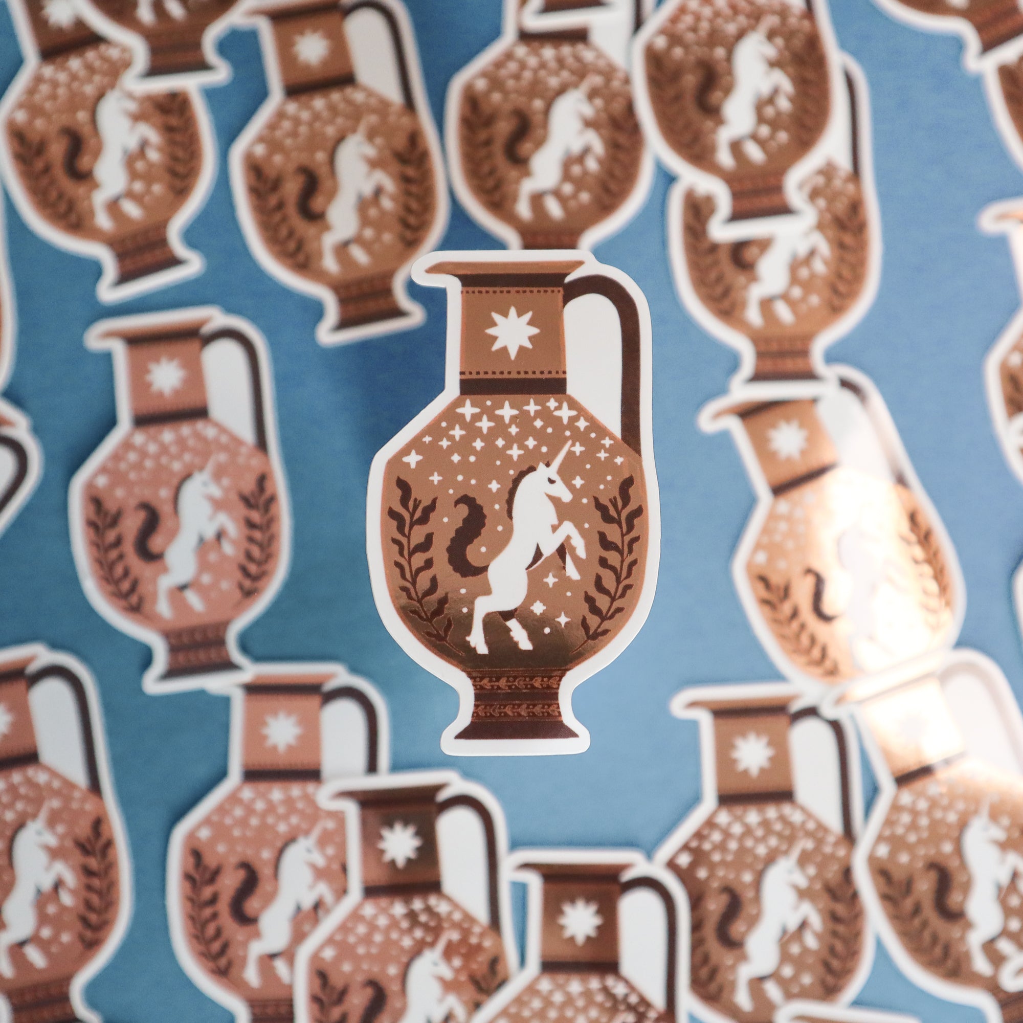 Papio Press Unicorn Vase Stickers, featuring a white unicorn with stars and leafy designs on brown vinyl, are scattered across a blue surface, with one sticker in the center foreground.