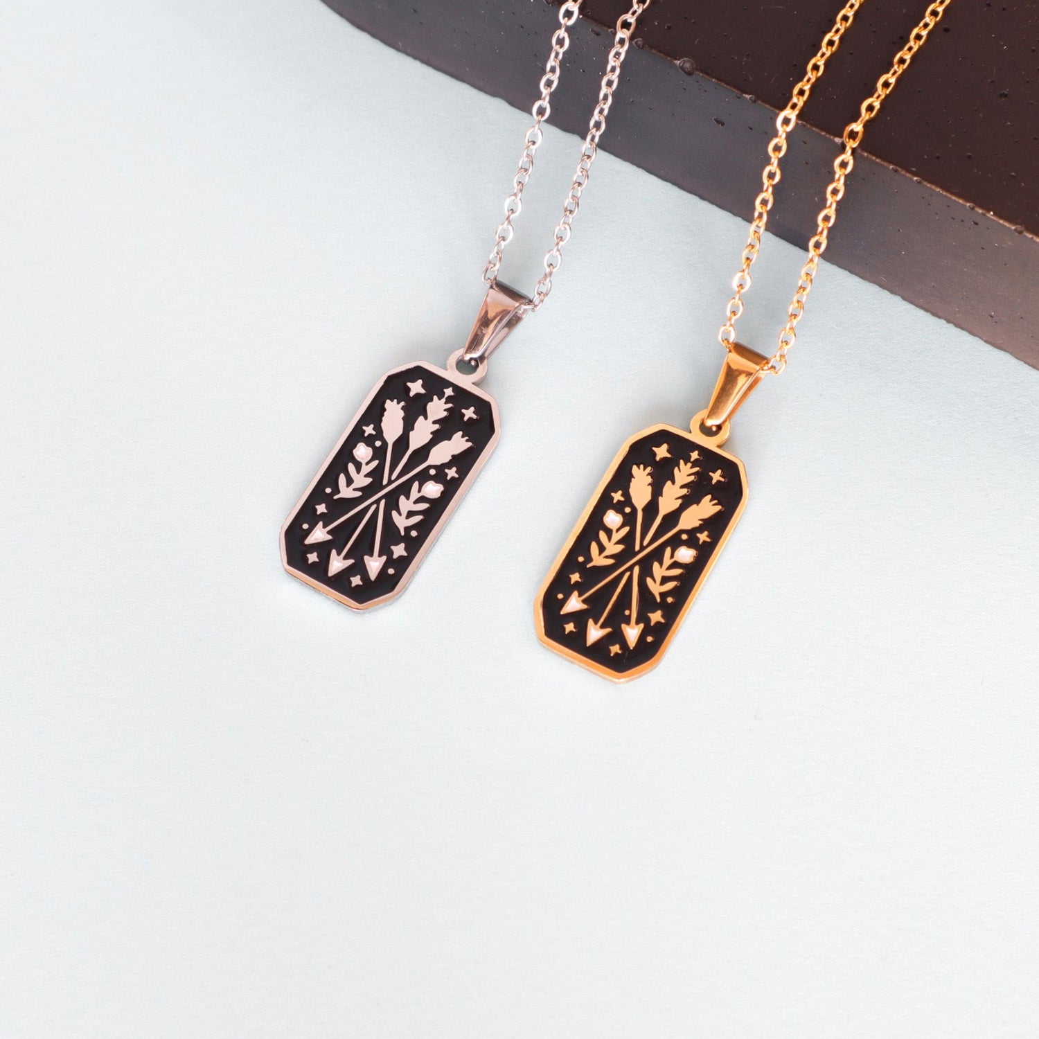 Two The Ranger Pendant Enamel Necklace pieces by Papio Press—one silver, one gold plated—feature rectangular floral enamel pendants on a white surface. Ideal statement jewelry for fantasy lovers seeking something unique.