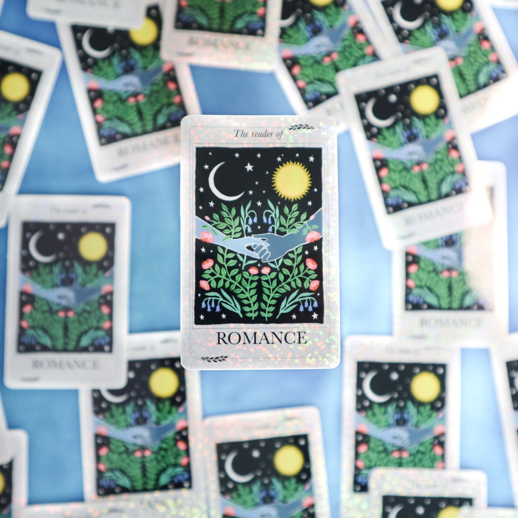 The Reader of Romance Sticker by Papio Press shows a tarot card labeled ROMANCE with two hands, moon, sun, and plants. This vinyl sticker has silver glitter accents and UV protection for lasting shine.
