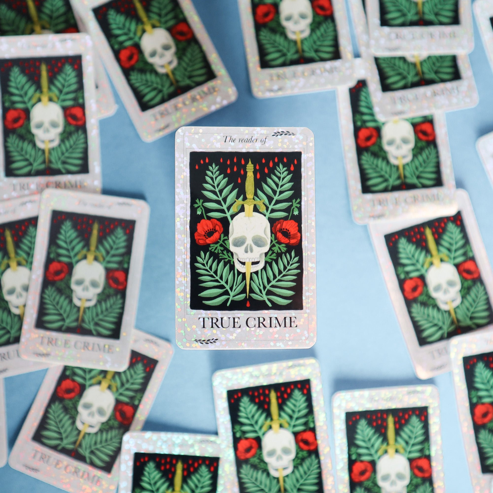 The Reader of True Crime Sticker by Papio Press shows a skull, green leaves, red flowers, and a dagger. Similar glittery vinyl stickers from the brand are scattered on a blue surface in the background.