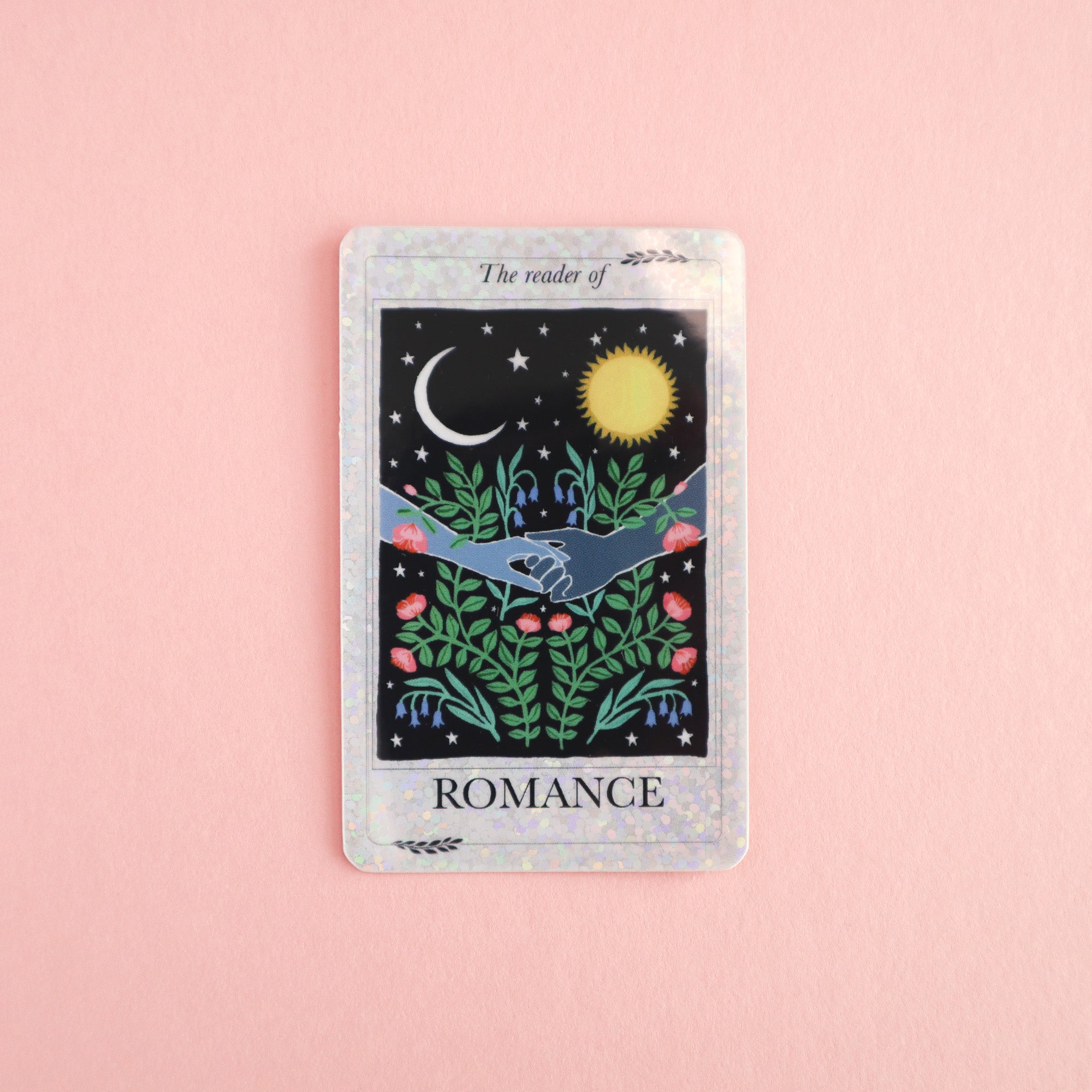 The Reader of Romance Sticker by Papio Press features two hands holding among plants, roses, a crescent moon, and a sun on a pink background. This vinyl sticker has an iridescent border and UV protection for lasting vibrance.