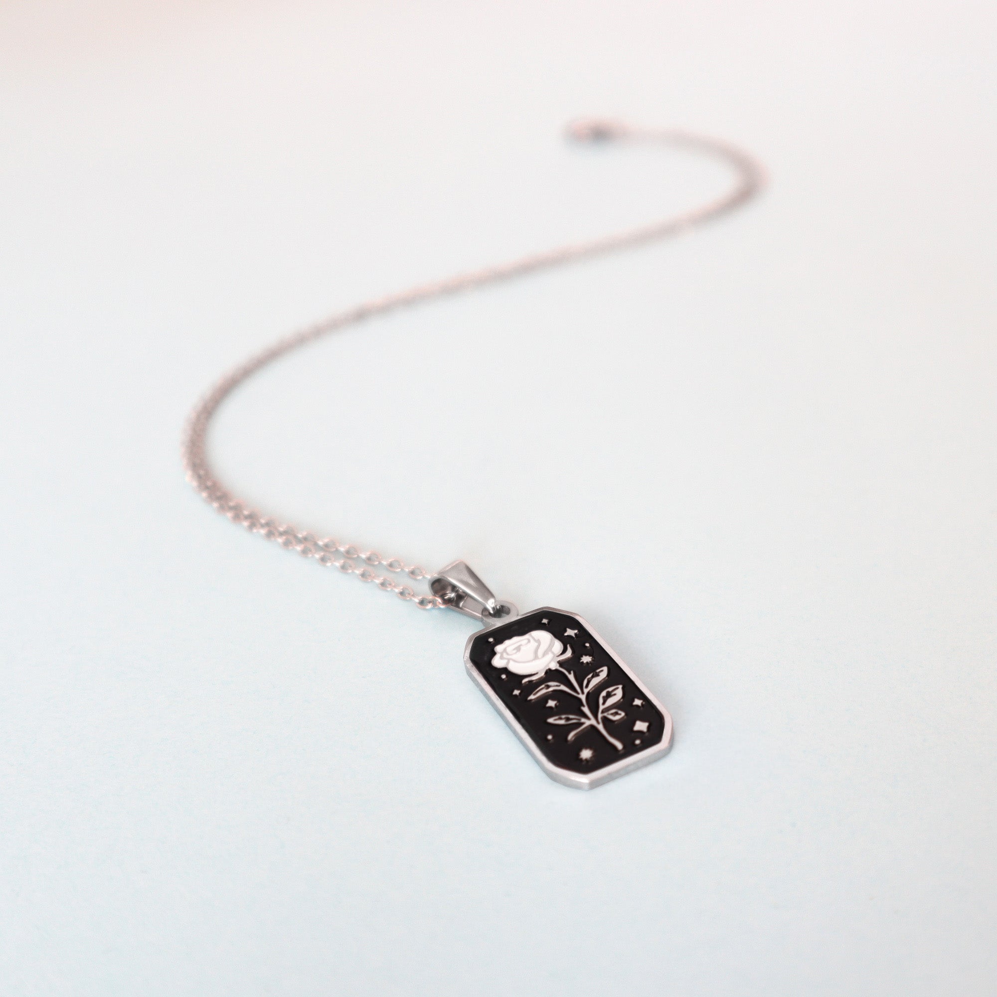 The Rose Pendant Enamel Necklace by Papio Press features a silver chain with a rectangular black pendant adorned with an engraved white rose and decorative accents, ideal for collectors or fantasy game fans, shown on a light background.
