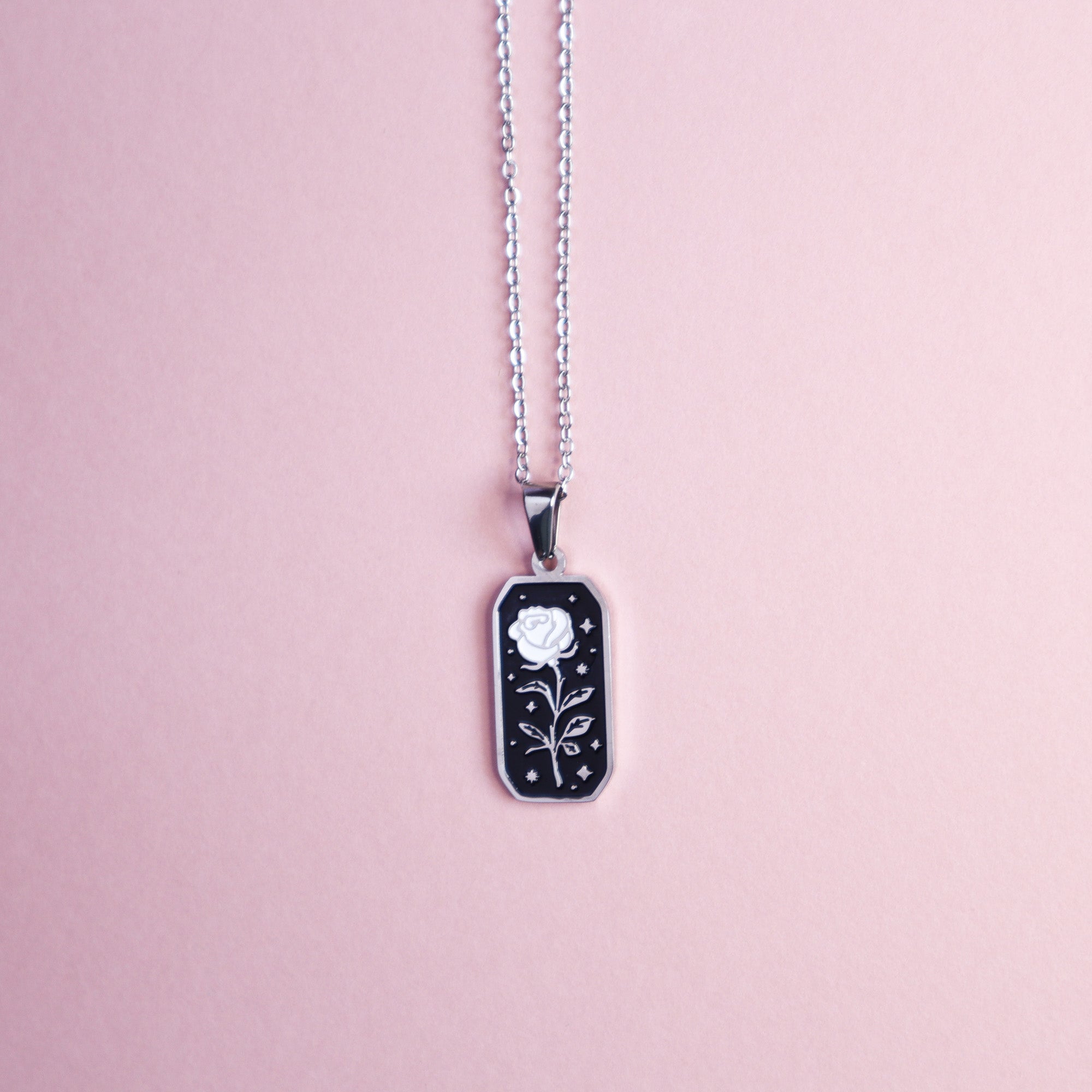 The Papio Press Rose Pendant Enamel Necklace features a rectangular black and silver design with a white rose and star accents, shown on a pastel pink background—ideal for rose jewelry collectors or fantasy game fans.