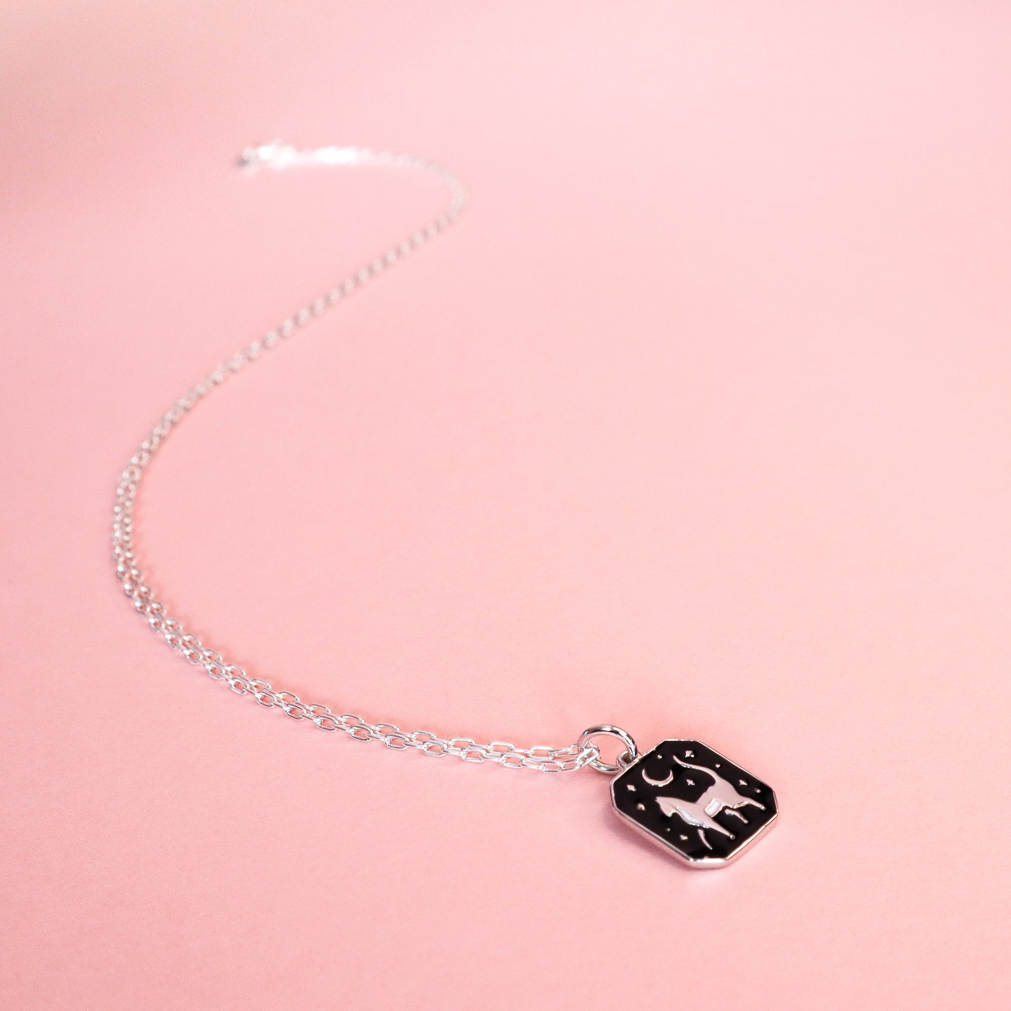 The Papio Press Silver Cat Pendant Enamel Necklace, featuring a rectangular black pendant with a white cat design, is displayed on a pastel pink background.