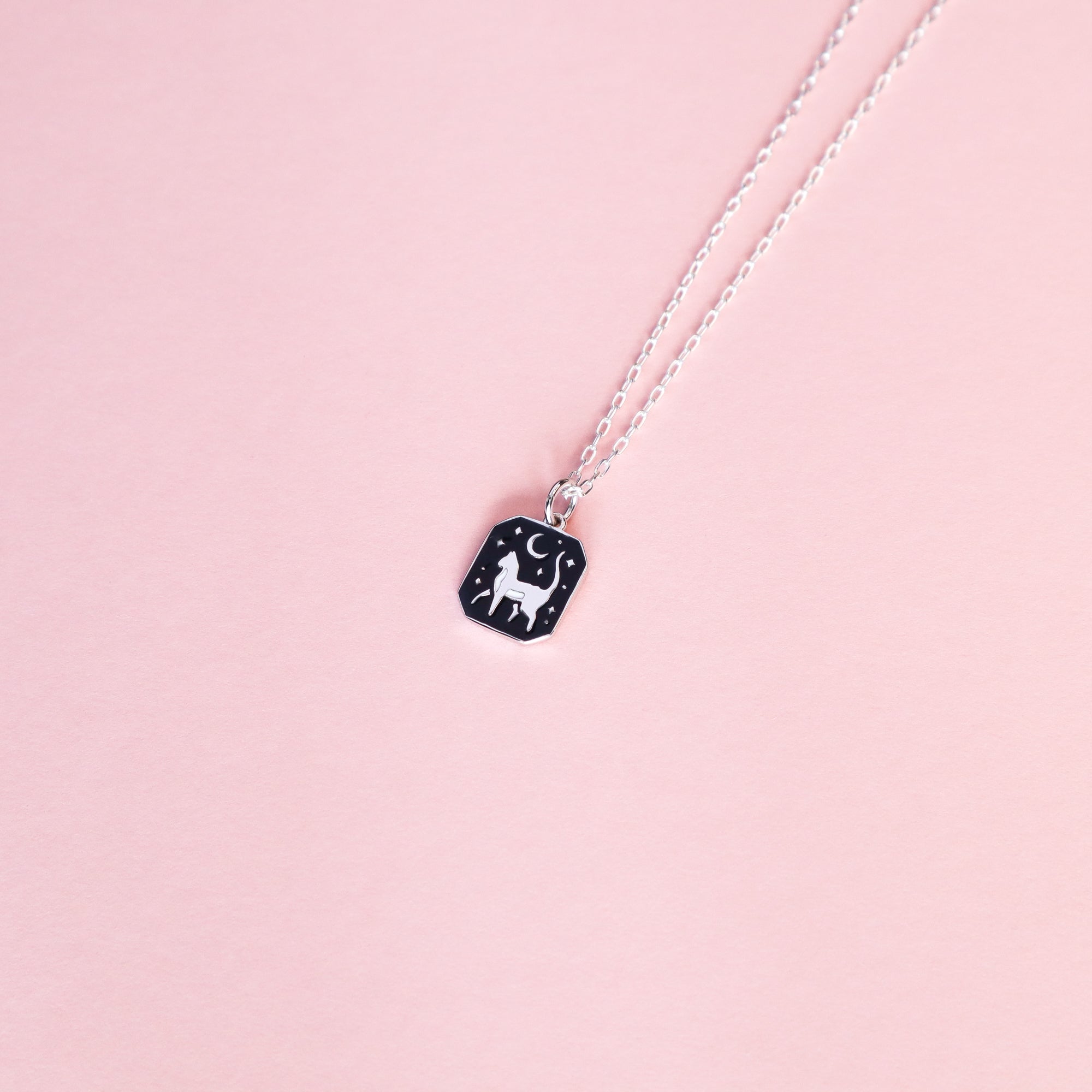 The Papio Press Silver Cat Pendant Enamel Necklace features a rectangular black pendant with a white cat, crescent moon, and stars, displayed on a pastel pink background. Made from recycled silver.
