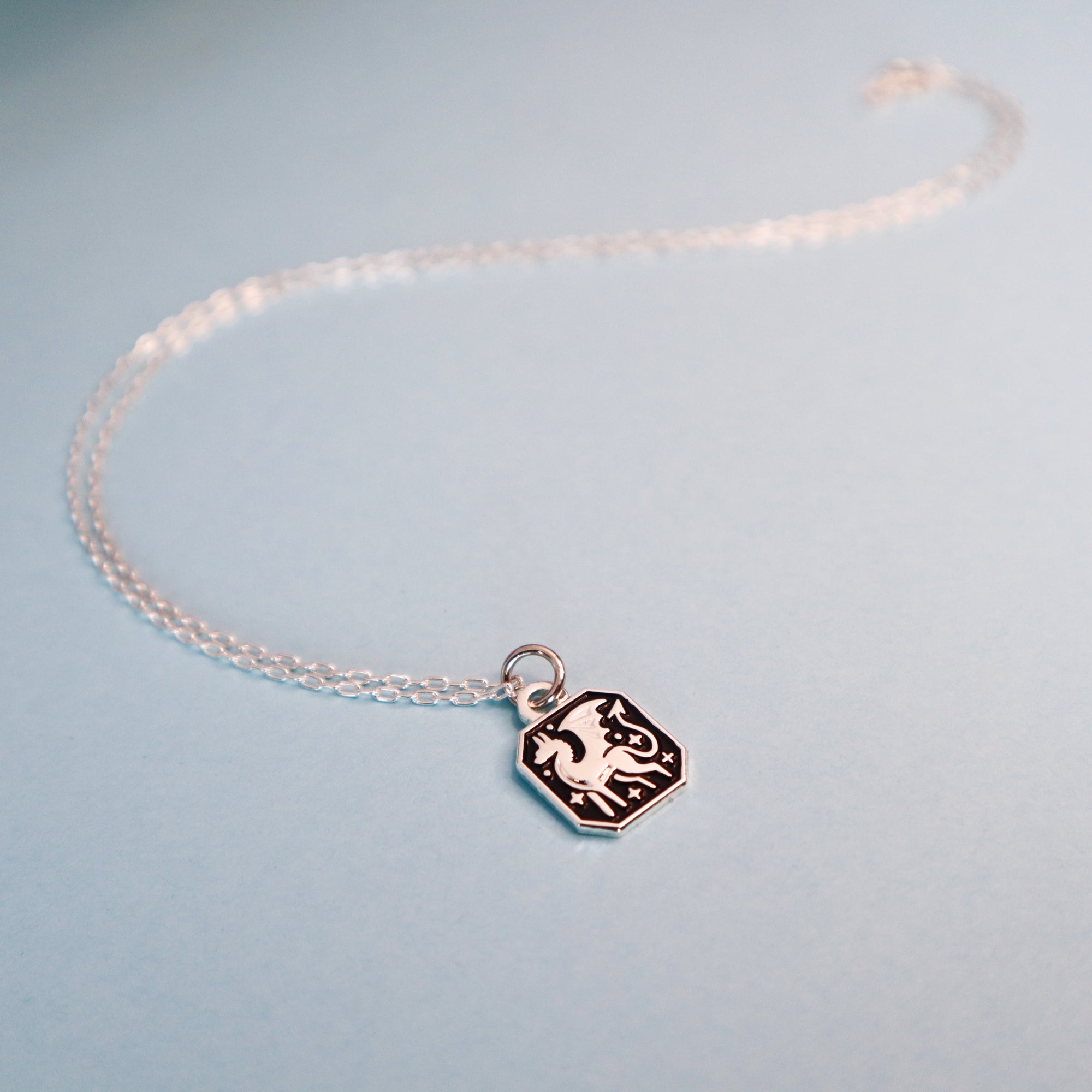 The Silver Dragon Pendant Enamel Necklace by Papio Press features a small, hexagonal black-and-silver recycled silver pendant with a white dragon design, shown on a light blue background.