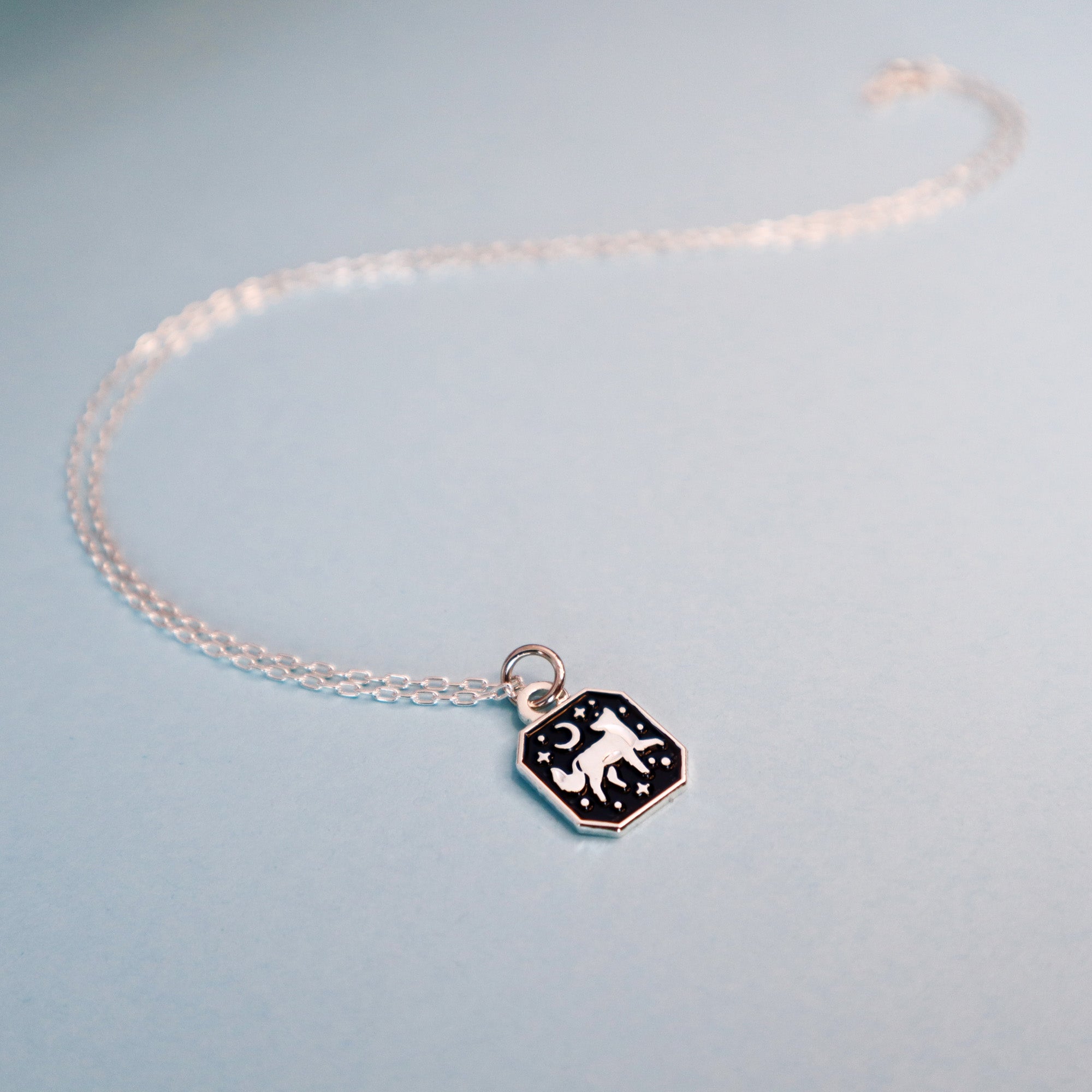 The Papio Press Silver Fox Pendant Enamel Necklace features a hexagonal recycled silver pendant with a fox, stars, and crescent moon on a black background, shown on a light blue surface.