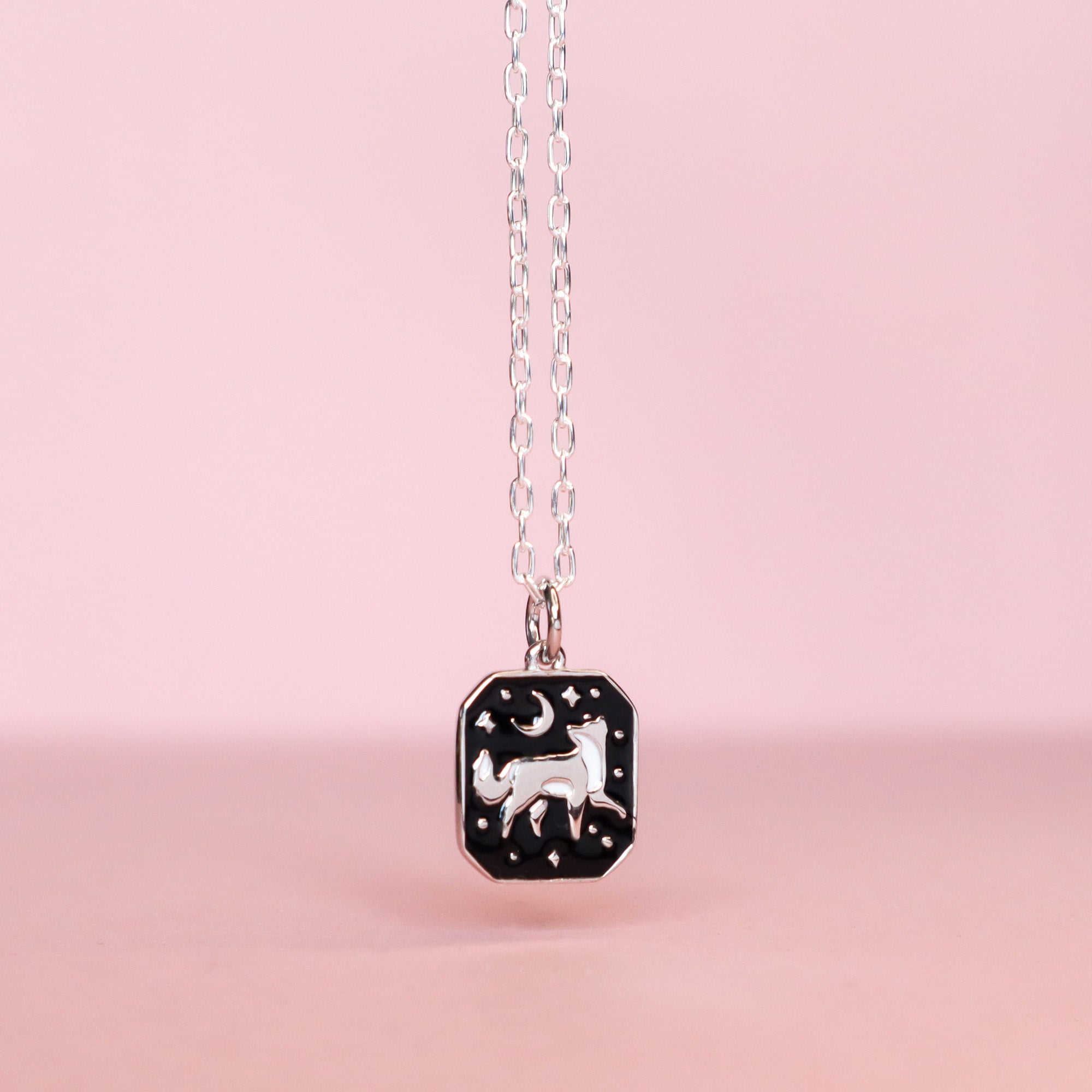 The Papio Press Silver Fox Pendant Enamel Necklace features a rectangular recycled silver pendant with a white fox, crescent moon, and stars on black enamel, set against a soft pink background.
