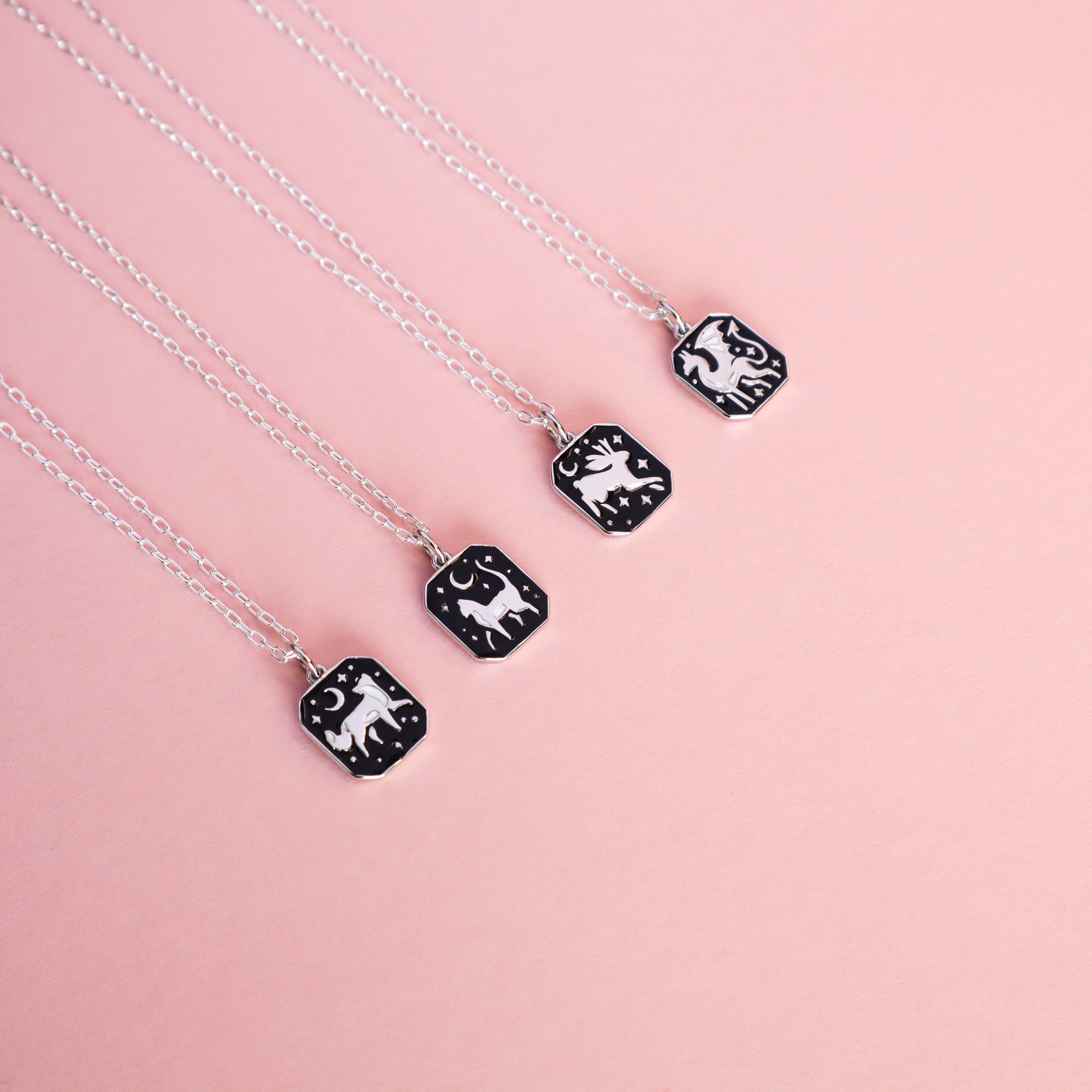 Four Papio Press Silver Fox Pendant Enamel Necklaces, made from recycled silver, feature rectangular black pendants with white animal and celestial designs. Arranged diagonally on light pink, each necklace blends elegance and artistry.