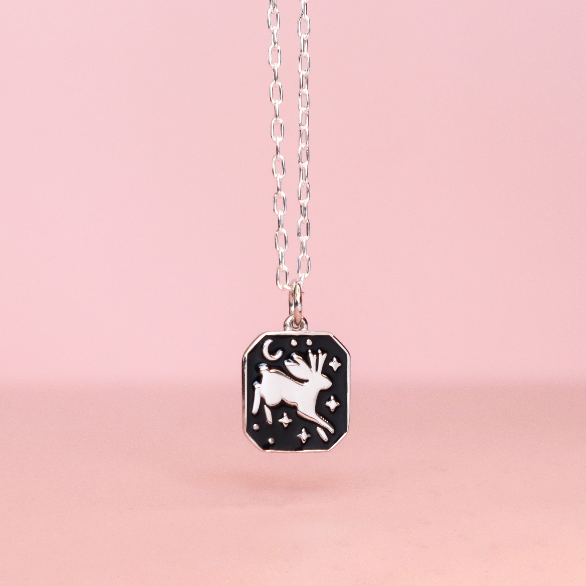 The Papio Press Silver Jackalope Pendant Enamel Necklace features a white jackalope, stars, and a crescent moon on black enamel, displayed against a soft pink background.