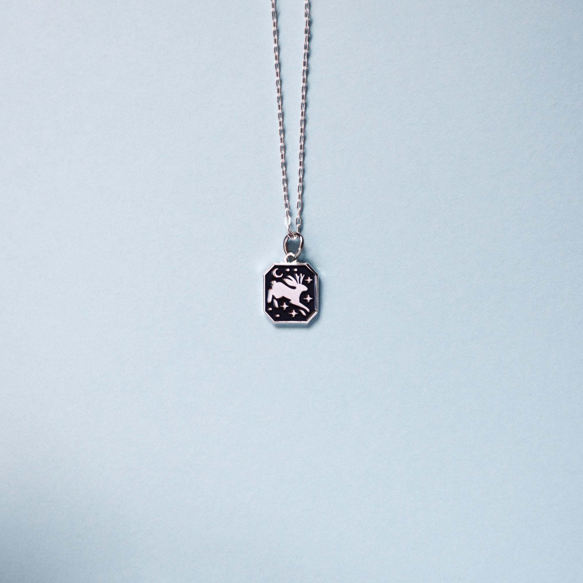 The Papio Press Silver Jackalope Pendant Enamel Necklace features a rectangular recycled silver pendant engraved with a deer, crescent moon, and stars, shown on a light blue background.