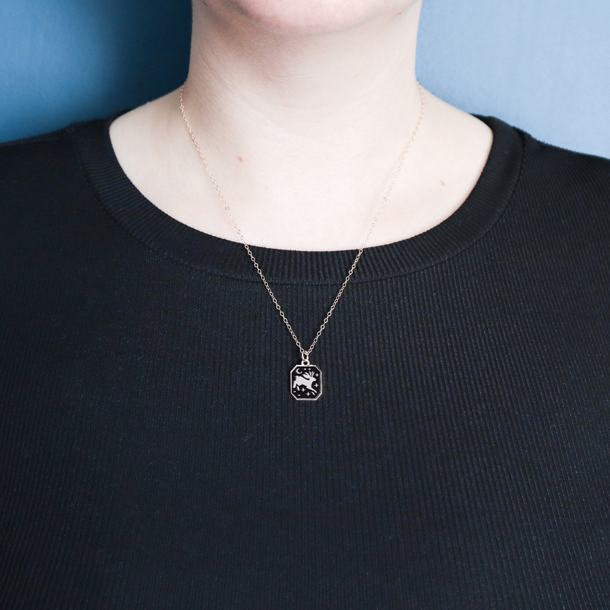 A person wearing a black ribbed shirt and a Papio Press Silver Jackalope Pendant Enamel Necklace stands against a blue background.