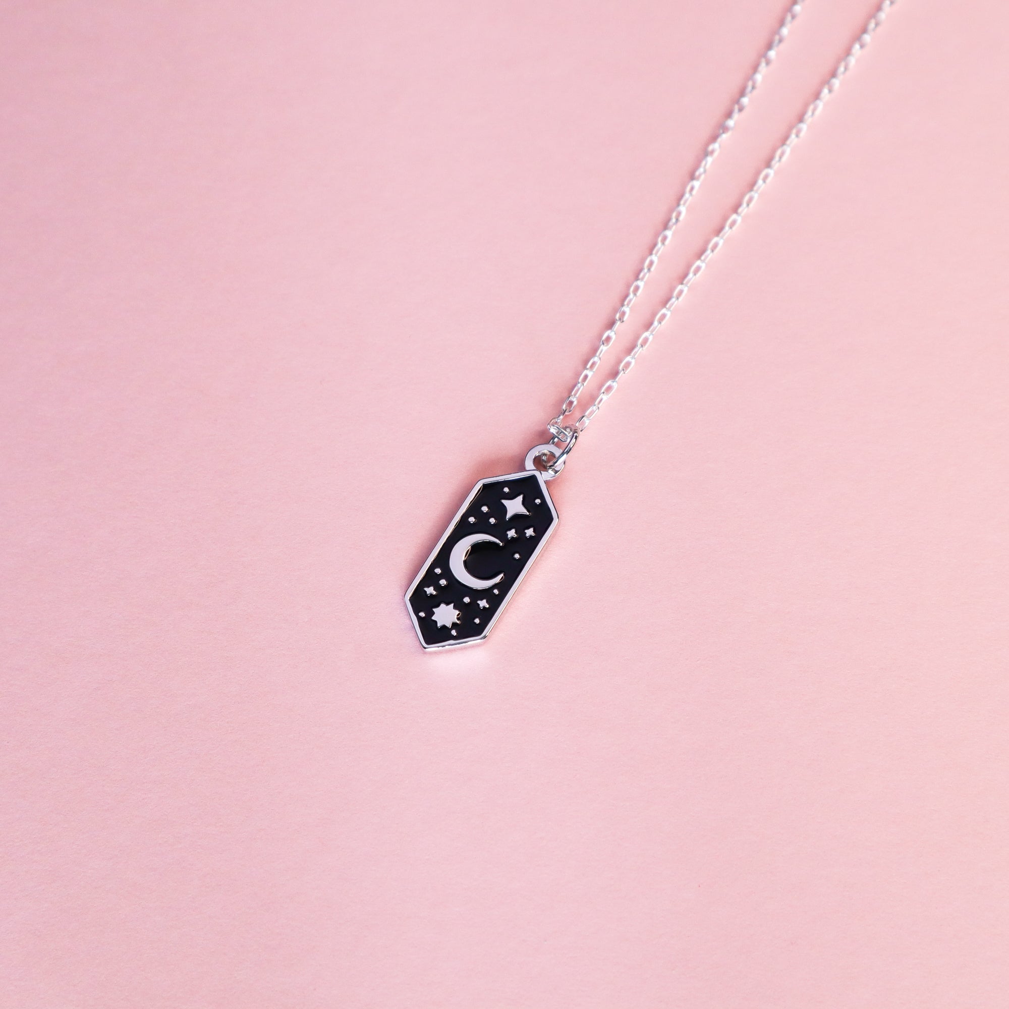 The Papio Press Silver Moon & Stars Enamel Necklace features a recycled silver chain and black hexagonal pendant with crescent moon and stars, shown on a light pink background—perfect for adding celestial charm to any outfit.