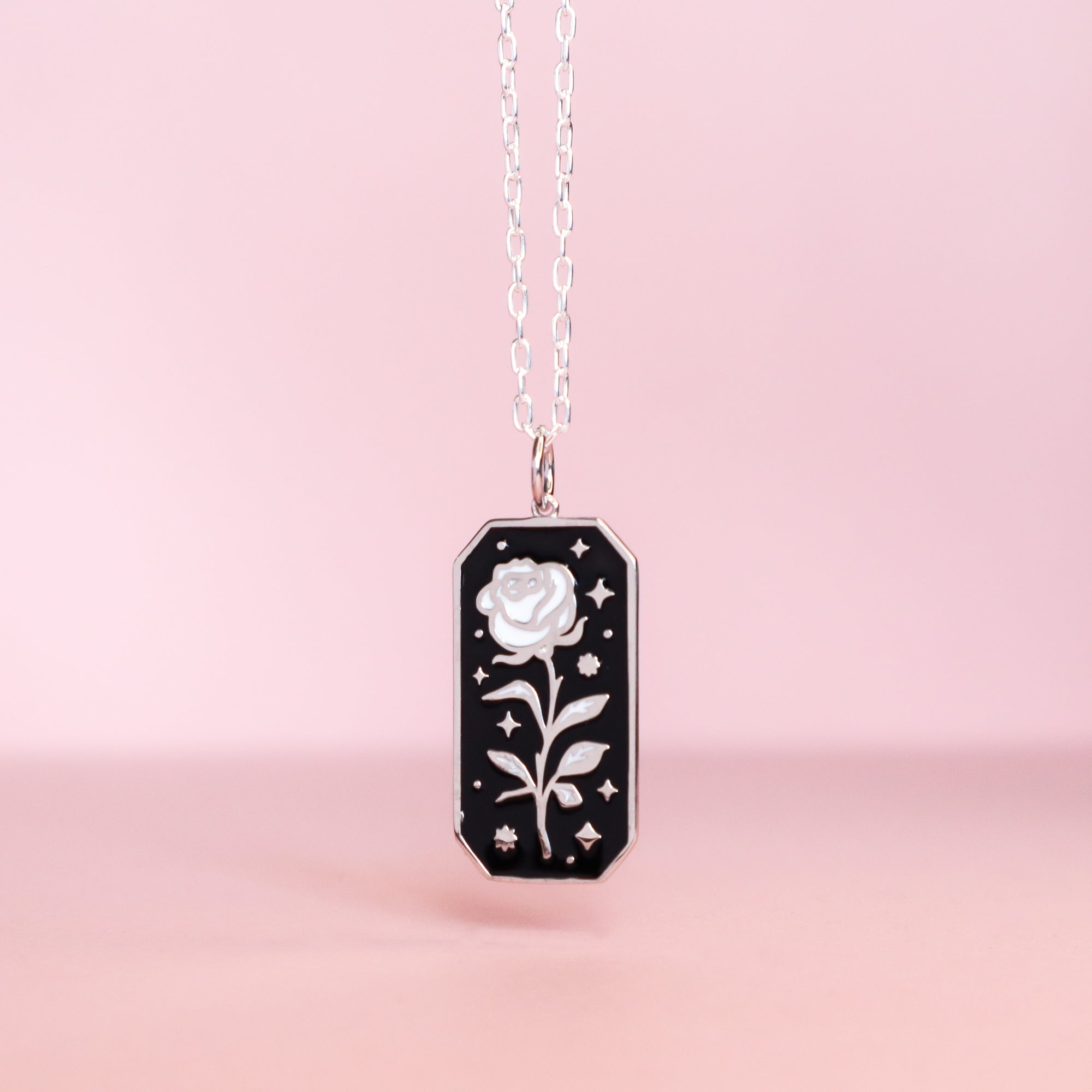 The Papio Press Silver Rose Enamel Necklace, made from recycled silver, features a rectangular black pendant with a white rose and star accents, shown against a soft pink background.