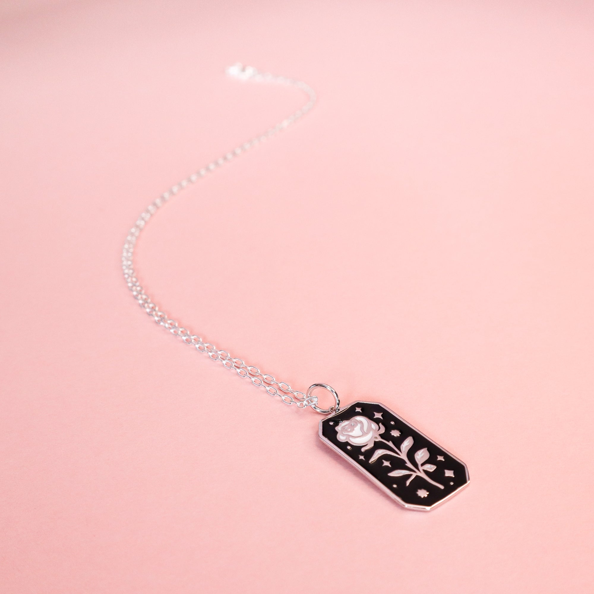 The Papio Press Silver Rose Enamel Necklace is crafted from recycled silver, featuring a rectangular black pendant with a silver rose and star accents on a light pink background.