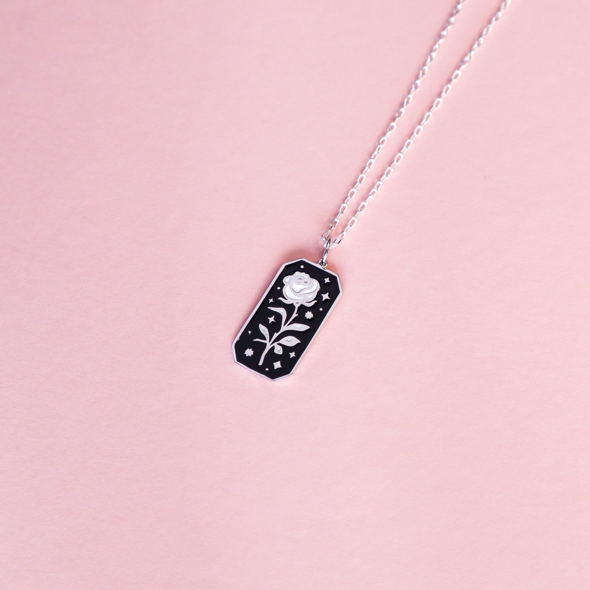 The Papio Press Silver Rose Enamel Necklace features a rectangular black pendant with a white rose and tiny stars, crafted from recycled silver and displayed on a light pink background.