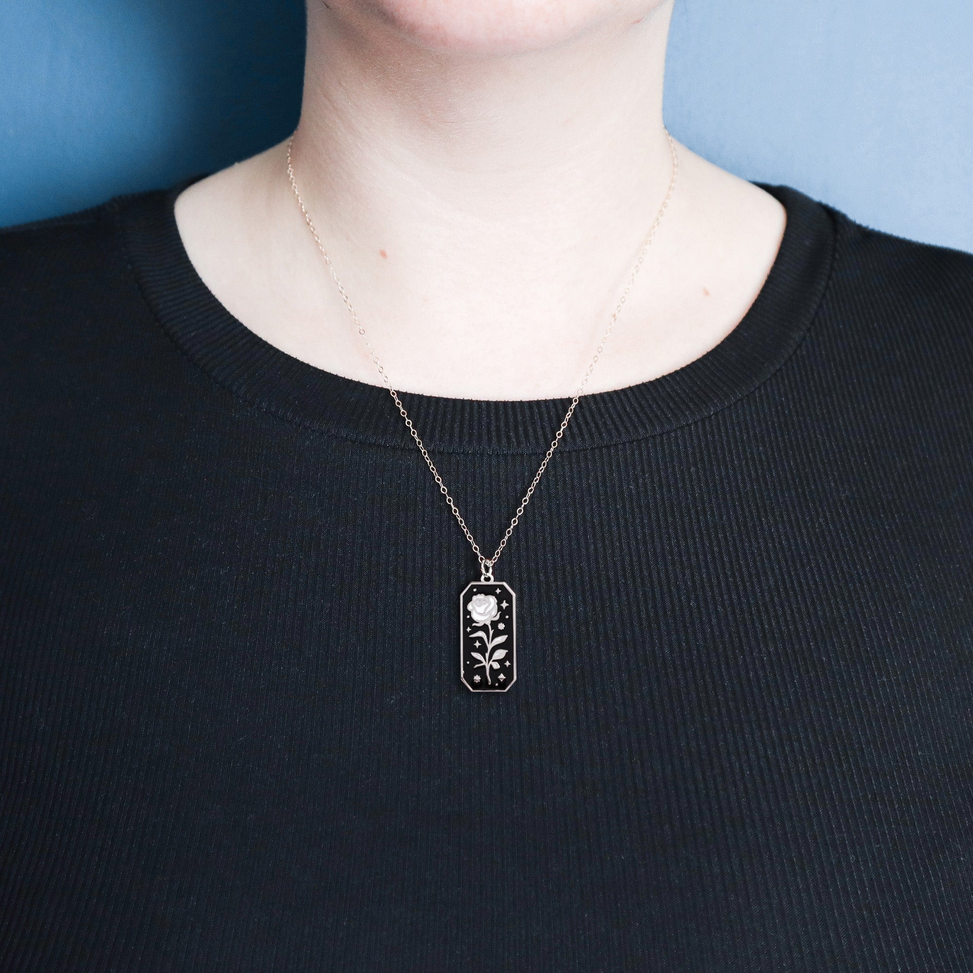 A person wears a Papio Press Silver Rose Enamel Necklace, featuring a floral design pendant, along with a black ribbed shirt and gold chain, standing against a blue background.