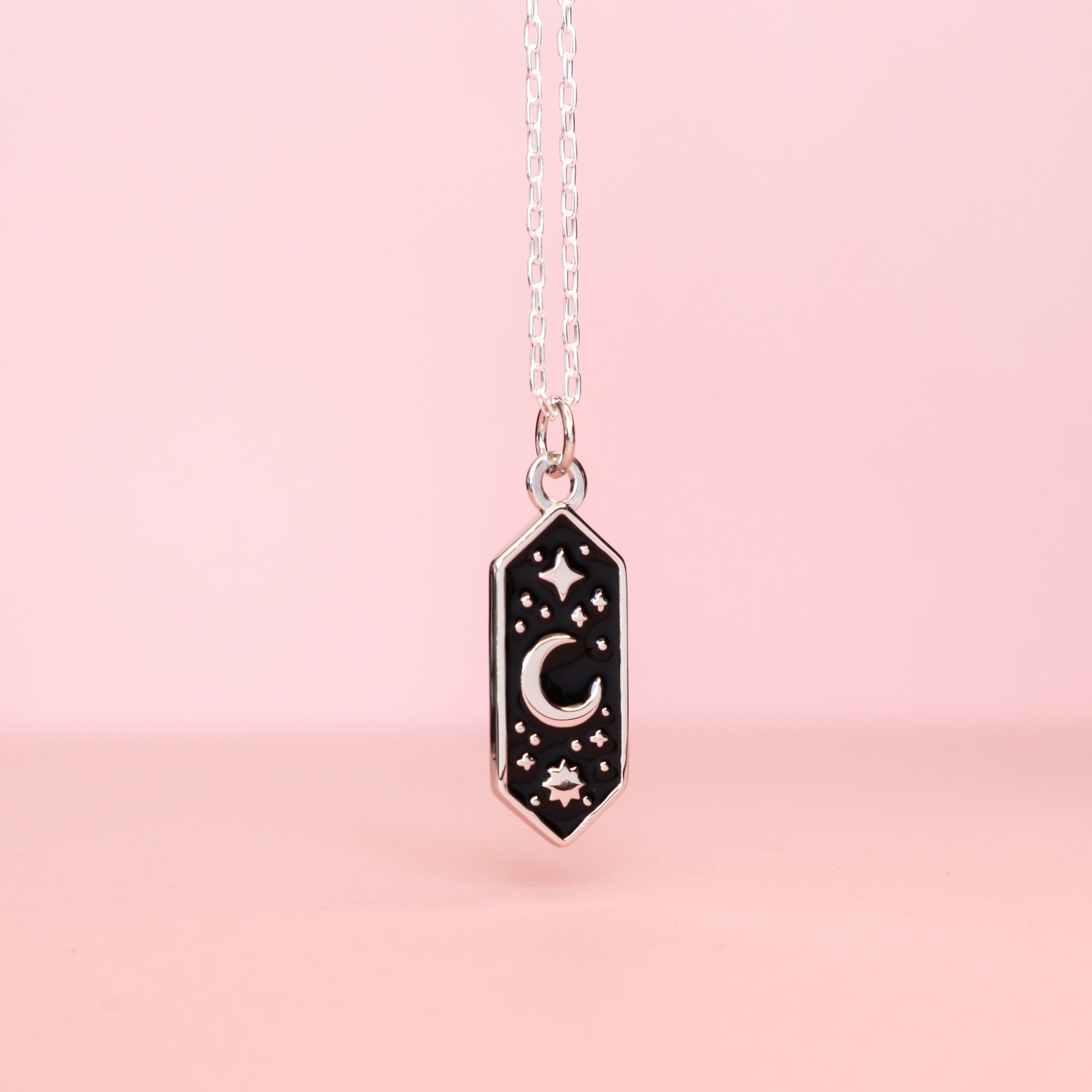 The Papio Press Silver Moon & Stars Enamel Necklace features a black hexagonal pendant with silver celestial details, showcased on a soft pink background.