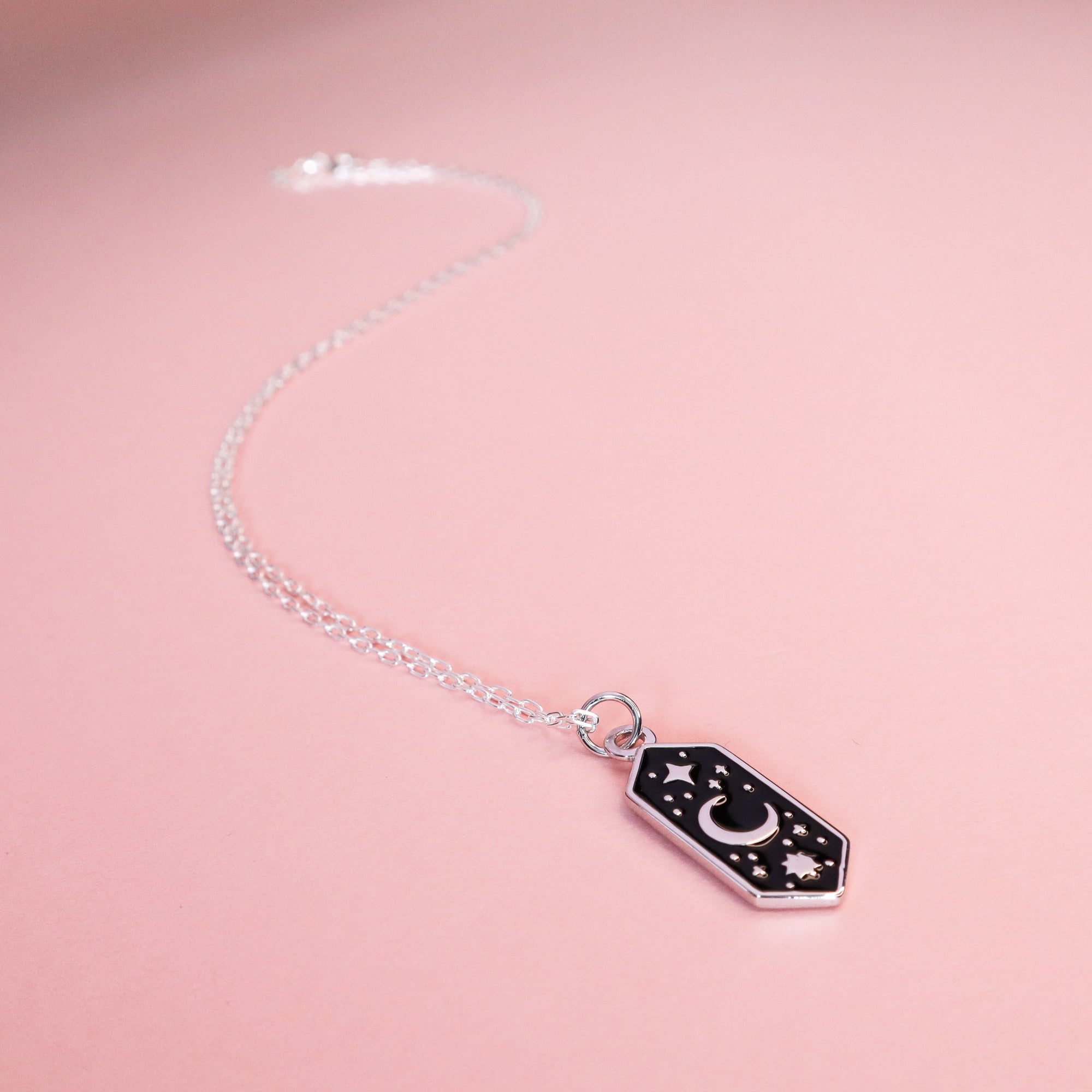The Papio Press Silver Moon & Stars Enamel Necklace features a hexagonal black enamel pendant with a crescent moon and stars, displayed on a light pink background.